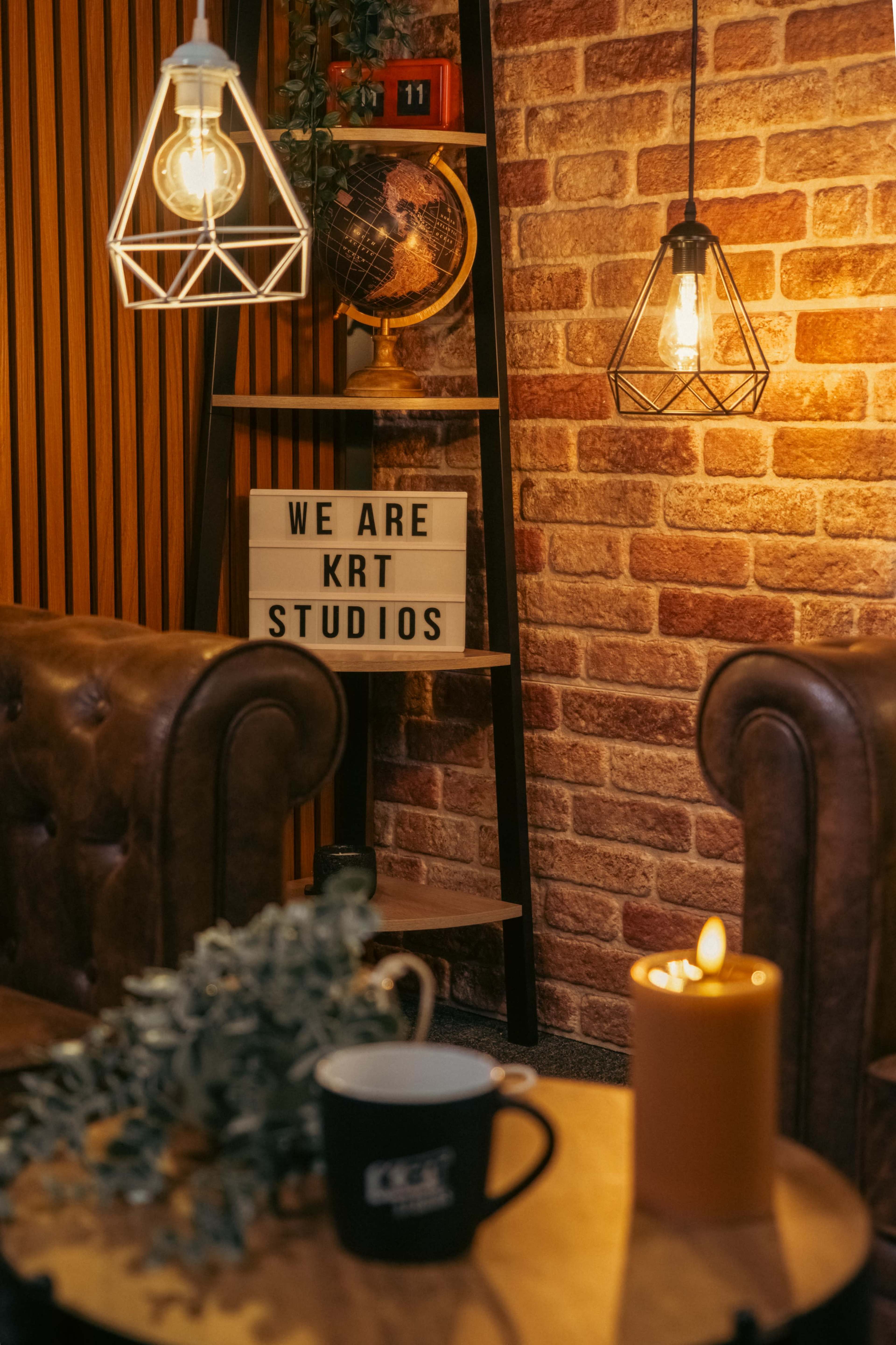 The image shows a cozy interior with industrial-style lighting, a sign that reads "WE ARE KRT STUDIOS," and a wooden table featuring a candle and a coffee cup.