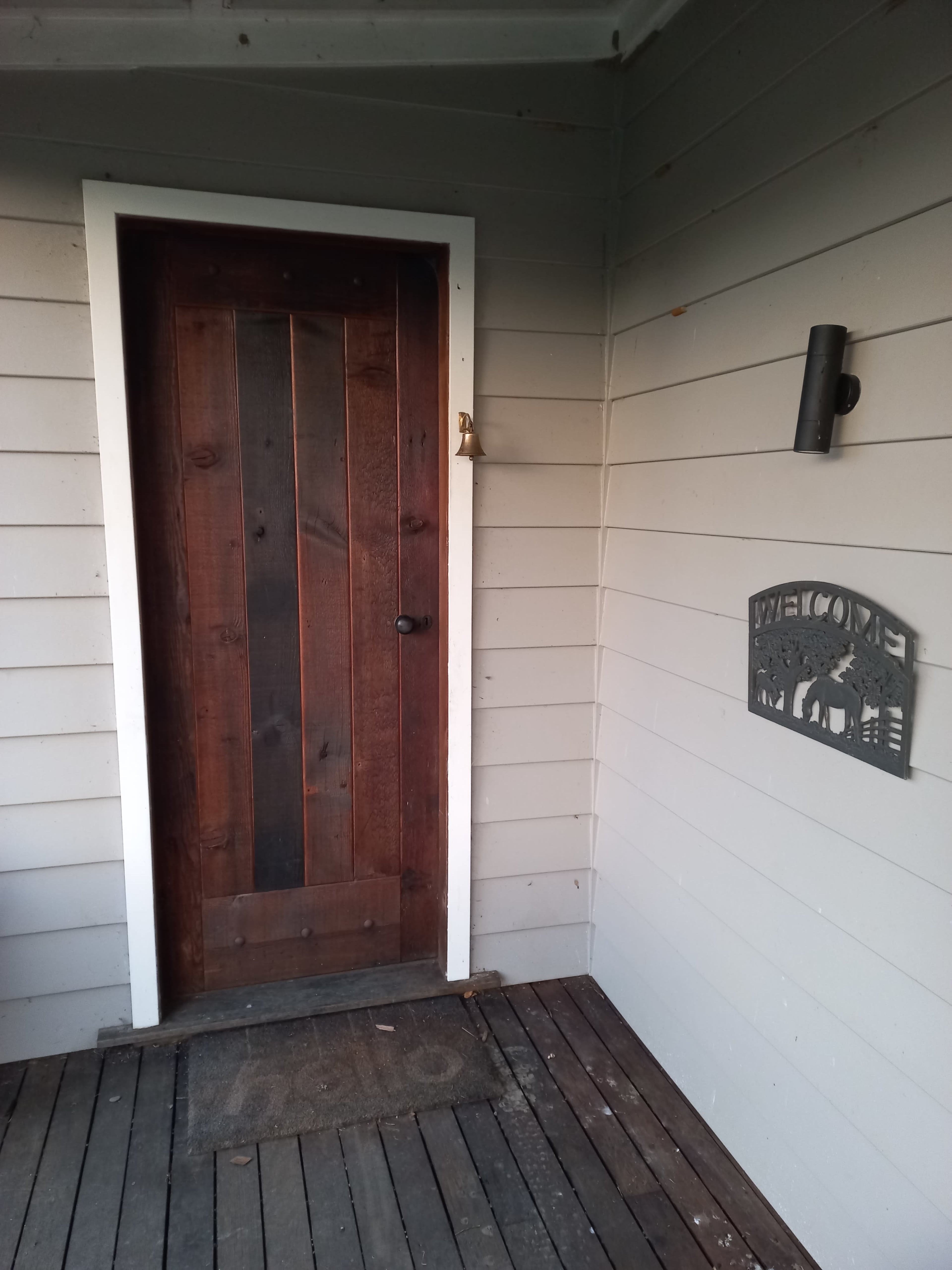 A wooden door with vertical planks, located on a porch with gray siding and a "Welcome" sign beside it.