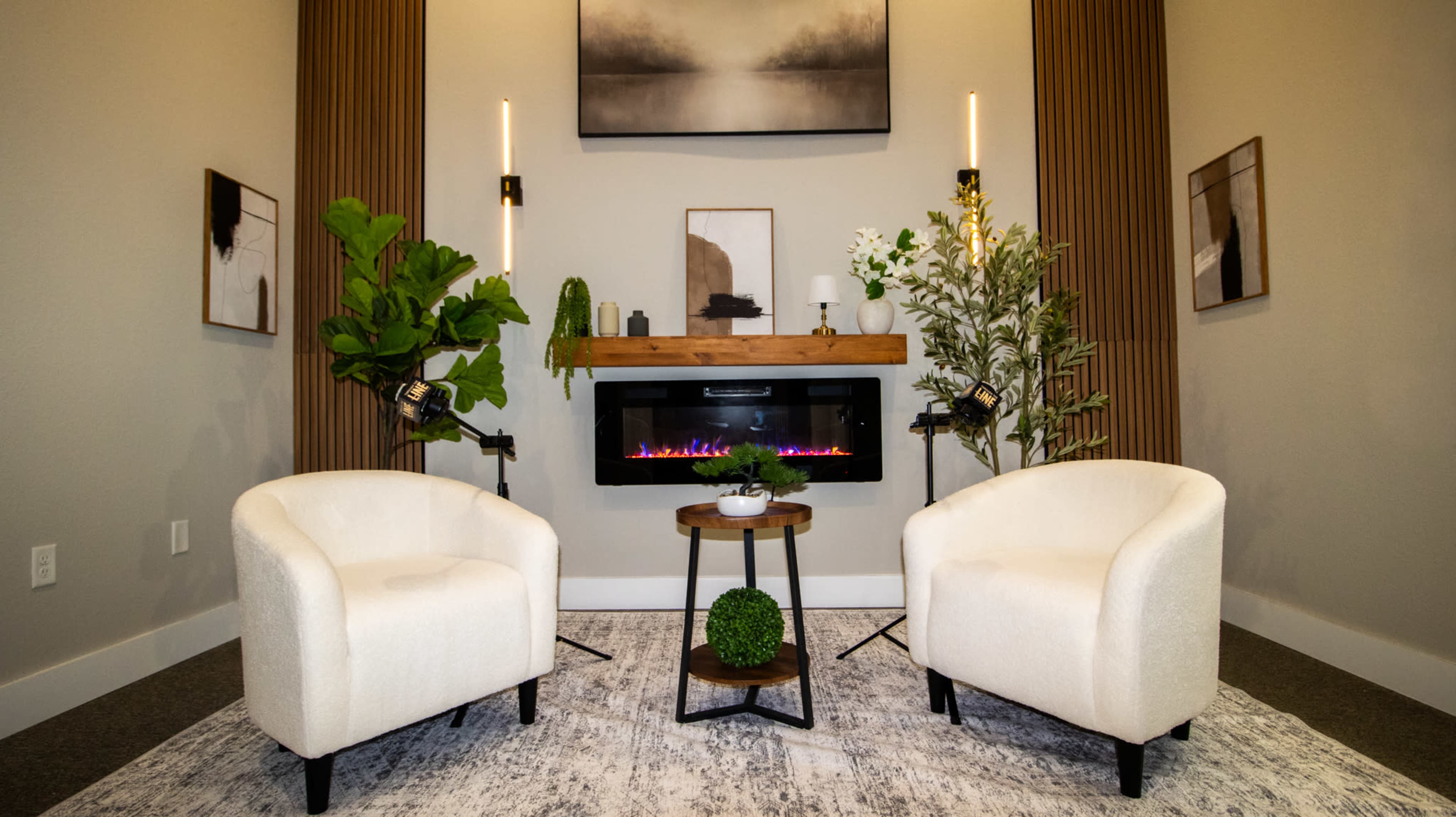 The image shows a cozy sitting area with two white armchairs surrounding a small table, an electric fireplace beneath a decorative shelf, and plants beside a neutral gray rug.