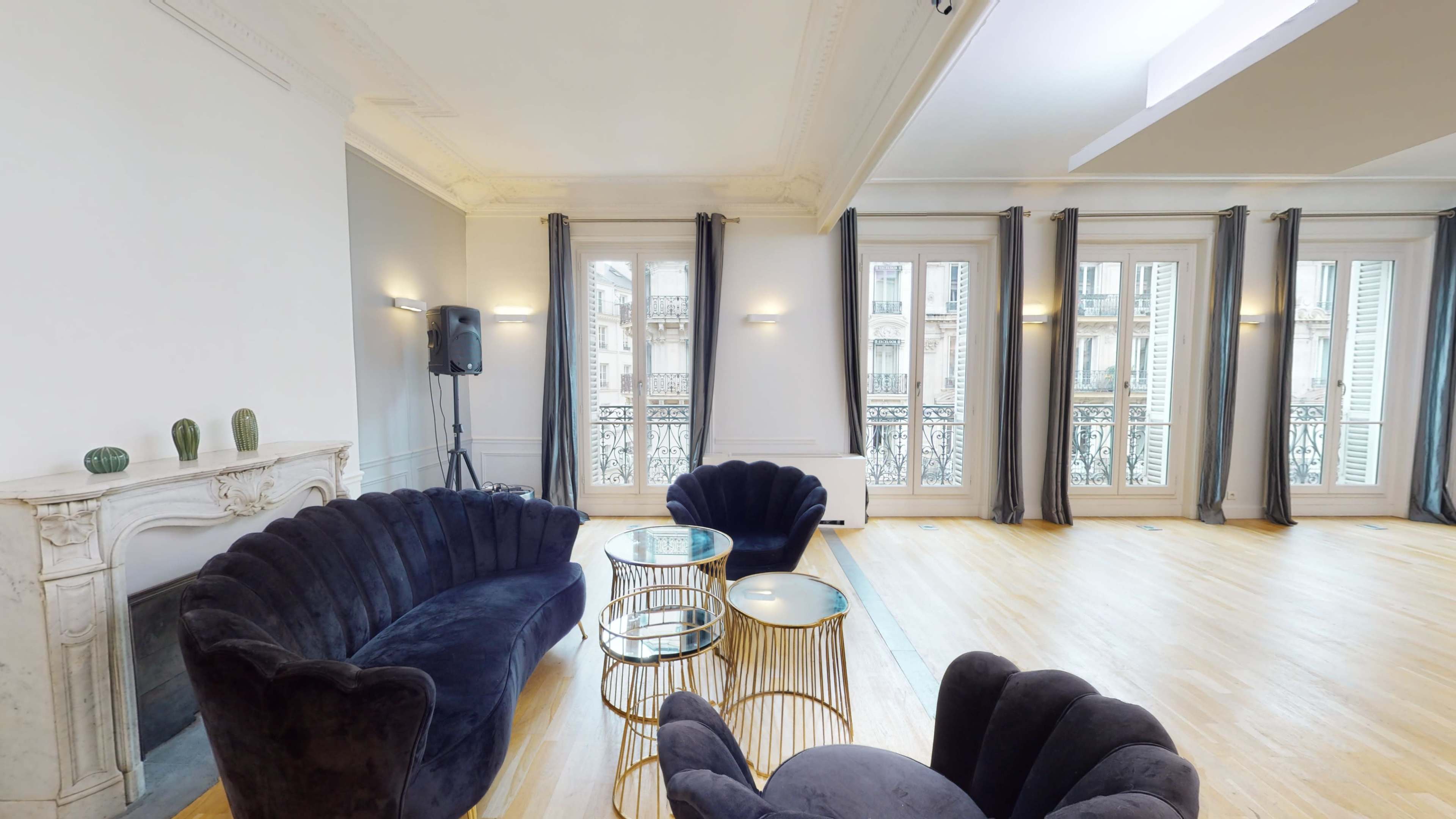 A spacious room featuring a dark velvet sofa, gold and glass side tables, and large windows with draped curtains.