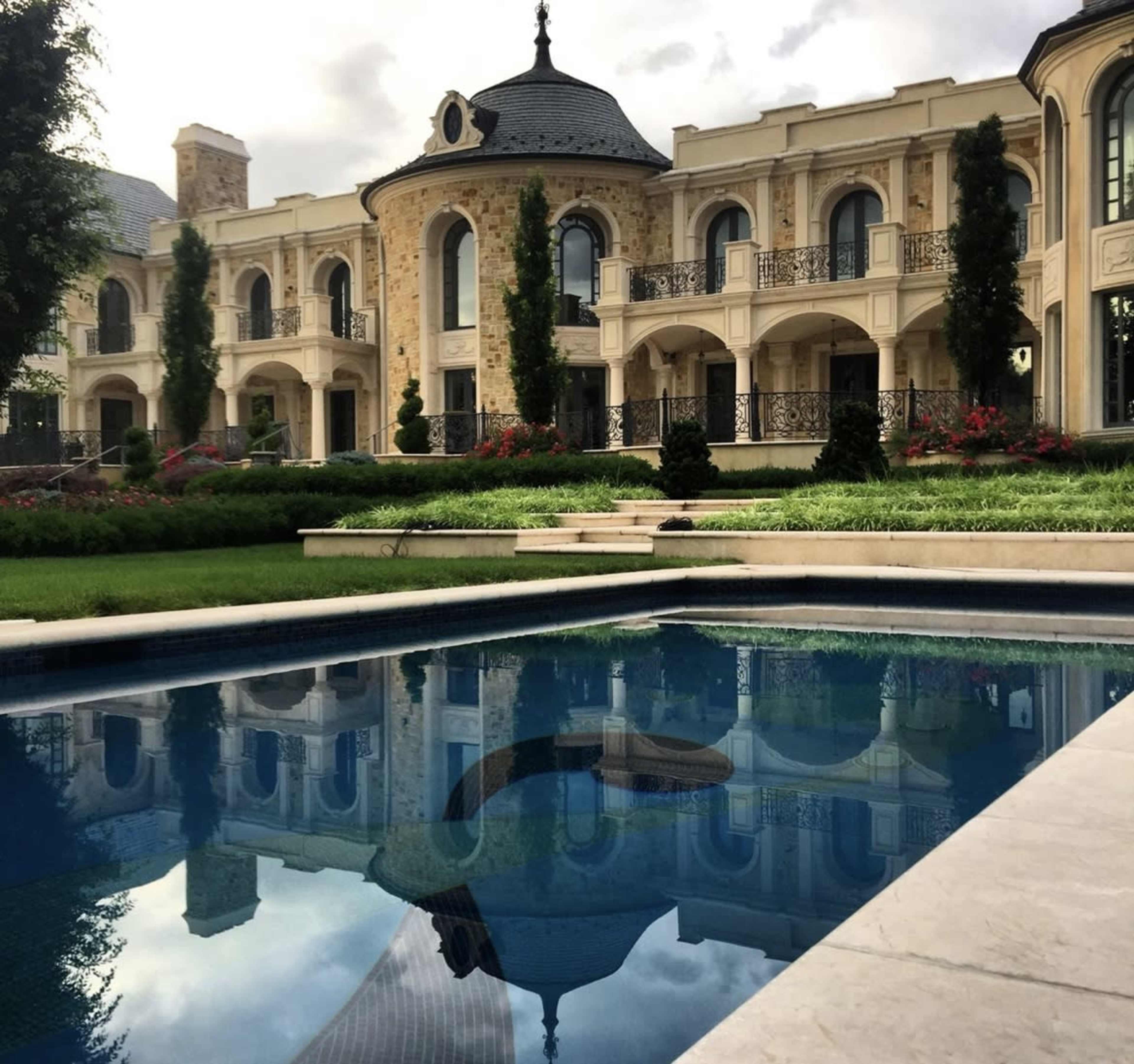 A large, elegant mansion with ornate architecture is reflected in a serene swimming pool, surrounded by lush landscaping and manicured gardens.