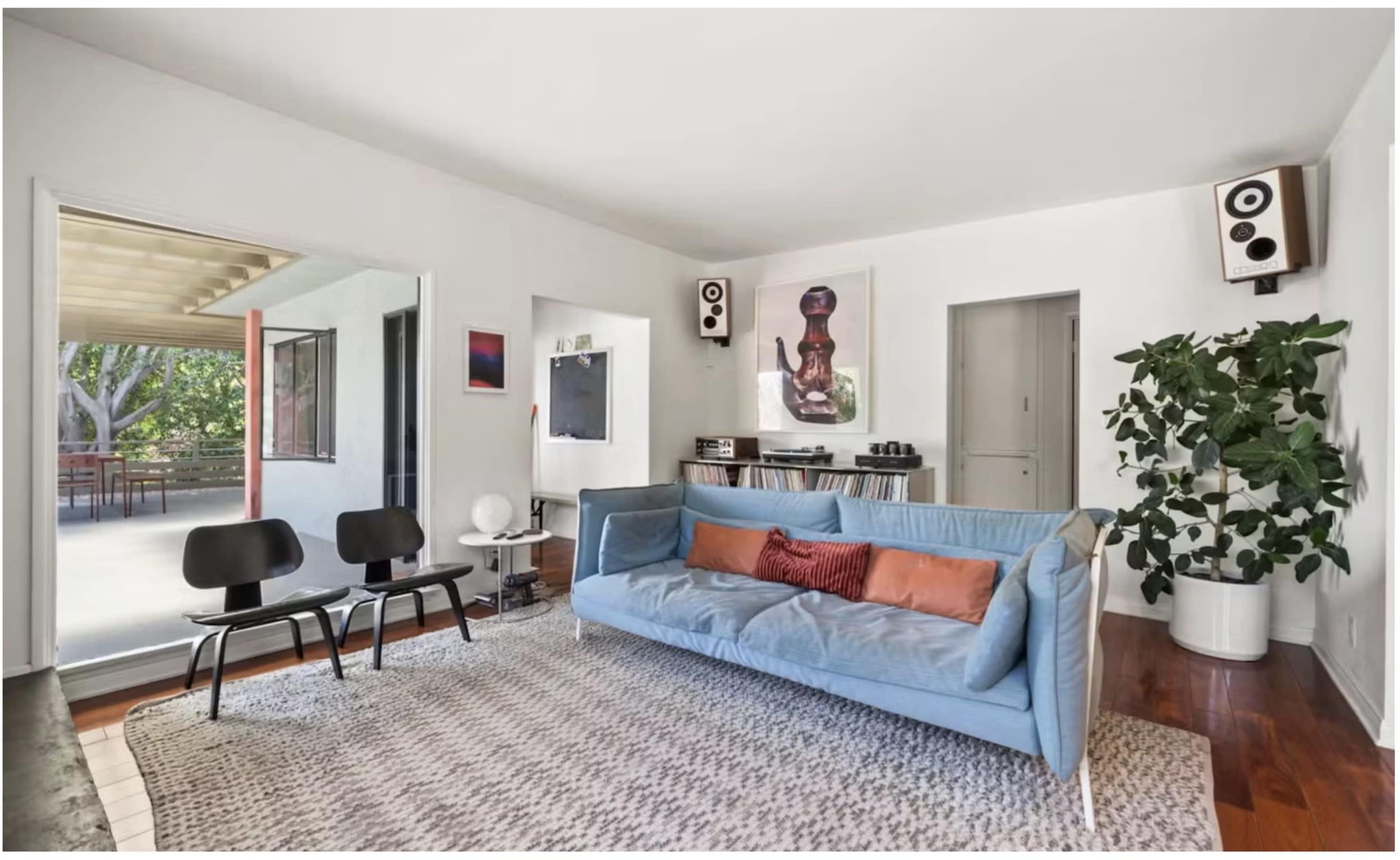 A modern living room featuring a blue sofa, two black chairs, a large area rug, and a plant, with natural light streaming in from adjacent outdoor space.