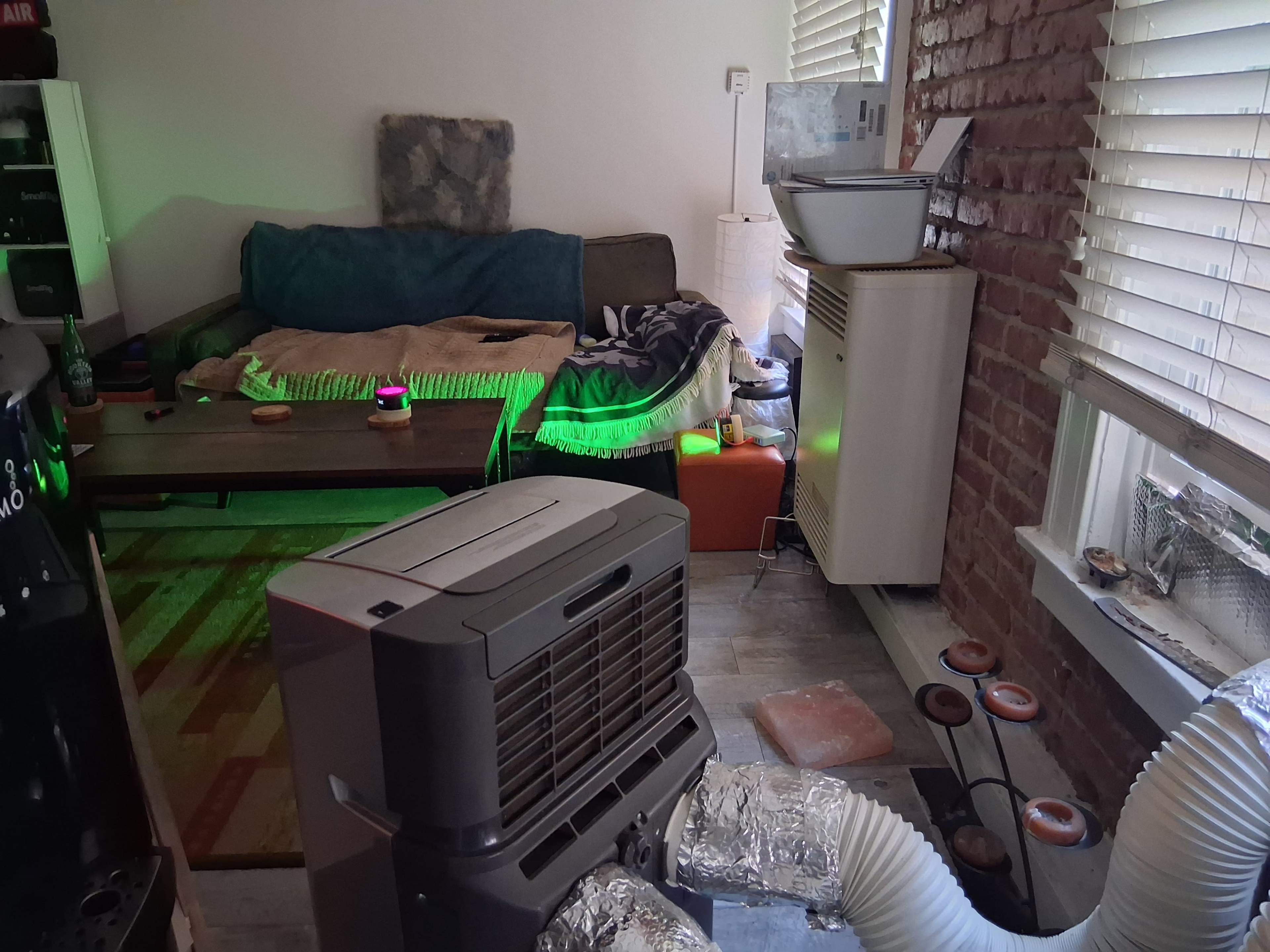 The image shows an indoor space with a couch, coffee table, portable air conditioner, and various household items, featuring exposed brick walls and window blinds.