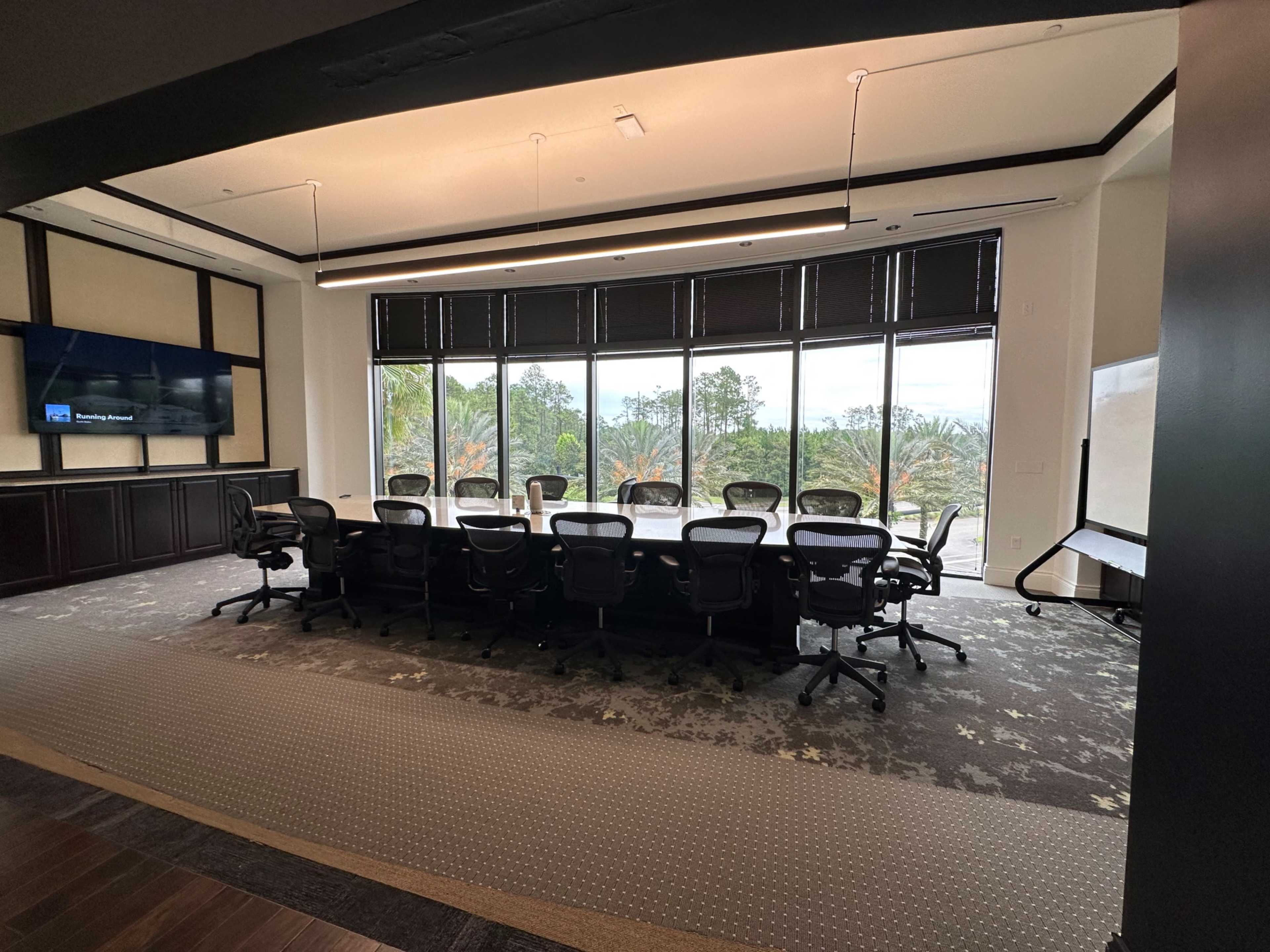 A large conference room features a long table surrounded by chairs, with windows displaying a view of greenery outside.