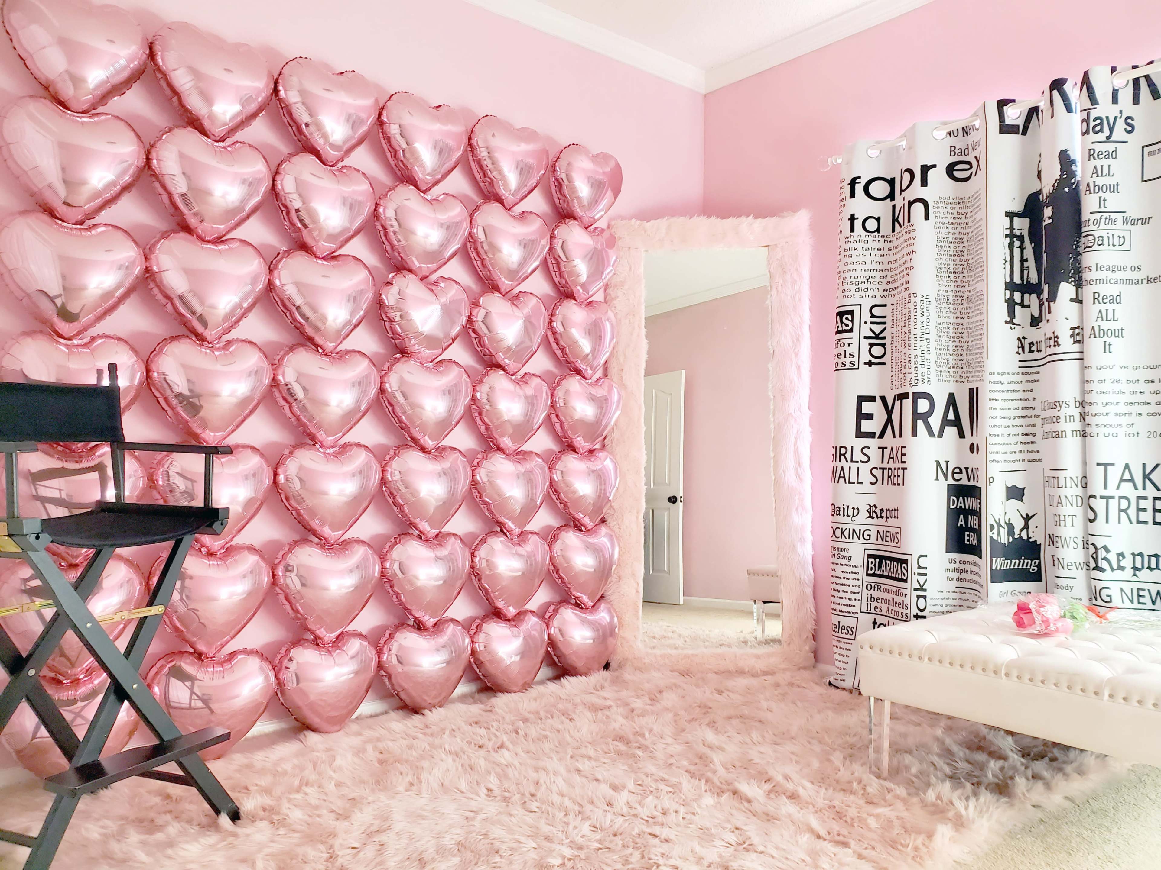A room features a wall covered in pink heart-shaped balloons, with a plush area rug and a decorative seating area.