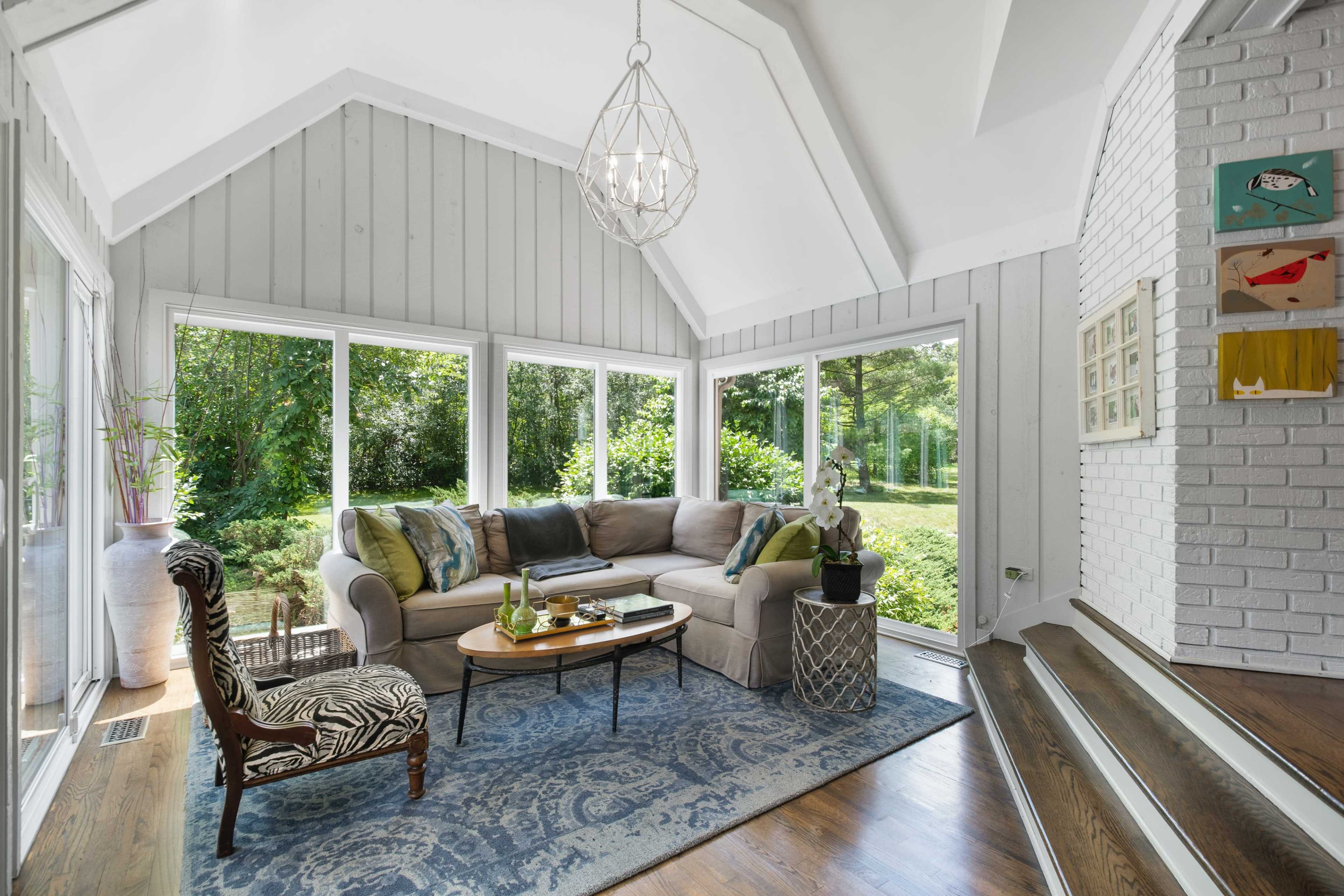 The image shows a sunlit sunroom featuring large windows, a sectional sofa, a round coffee table, and a decorative chair, with a view of a green garden outside.