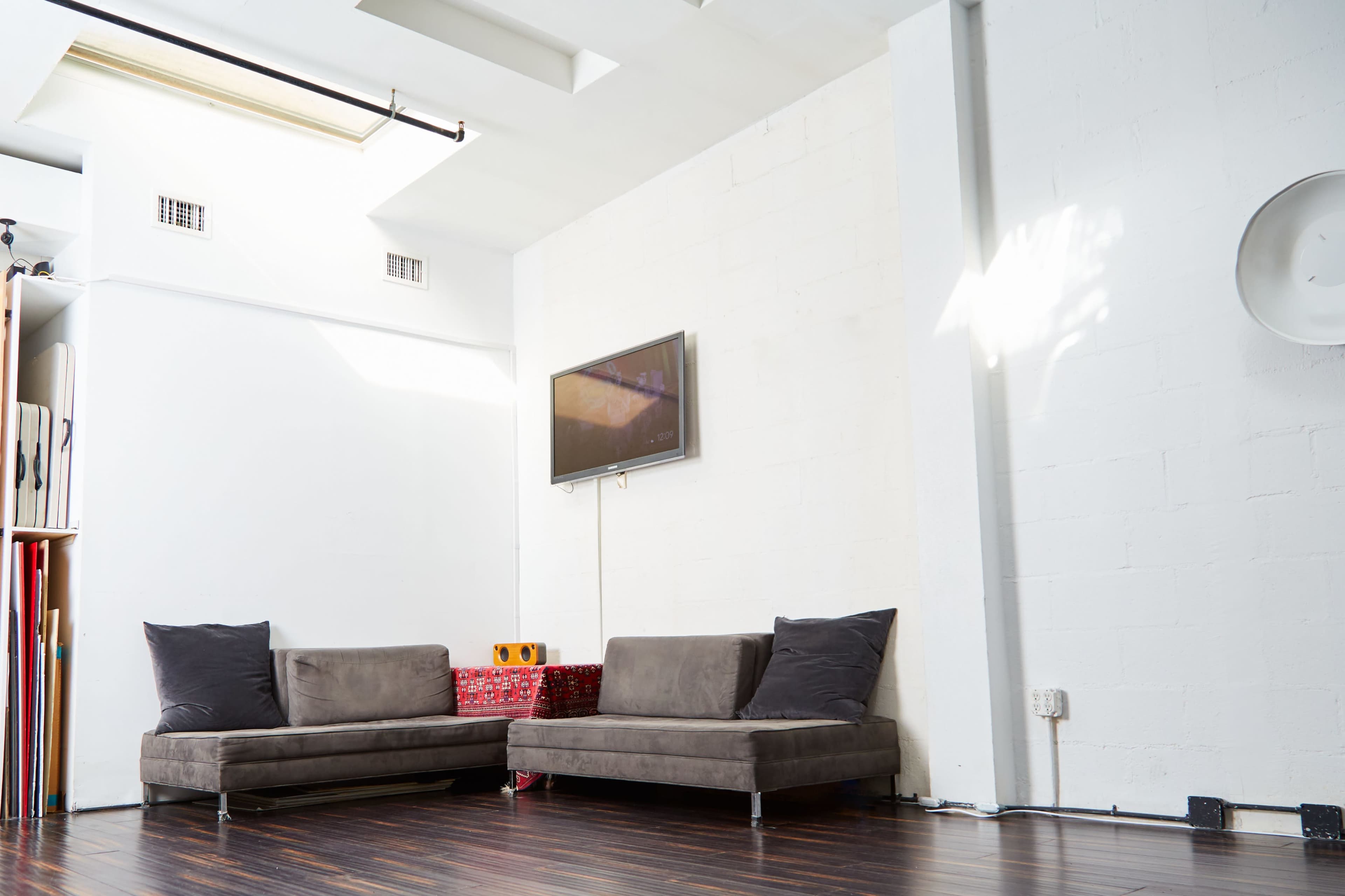 The image shows a minimalistic interior space featuring two gray couches, a wall-mounted television, and a white wall with wooden flooring.