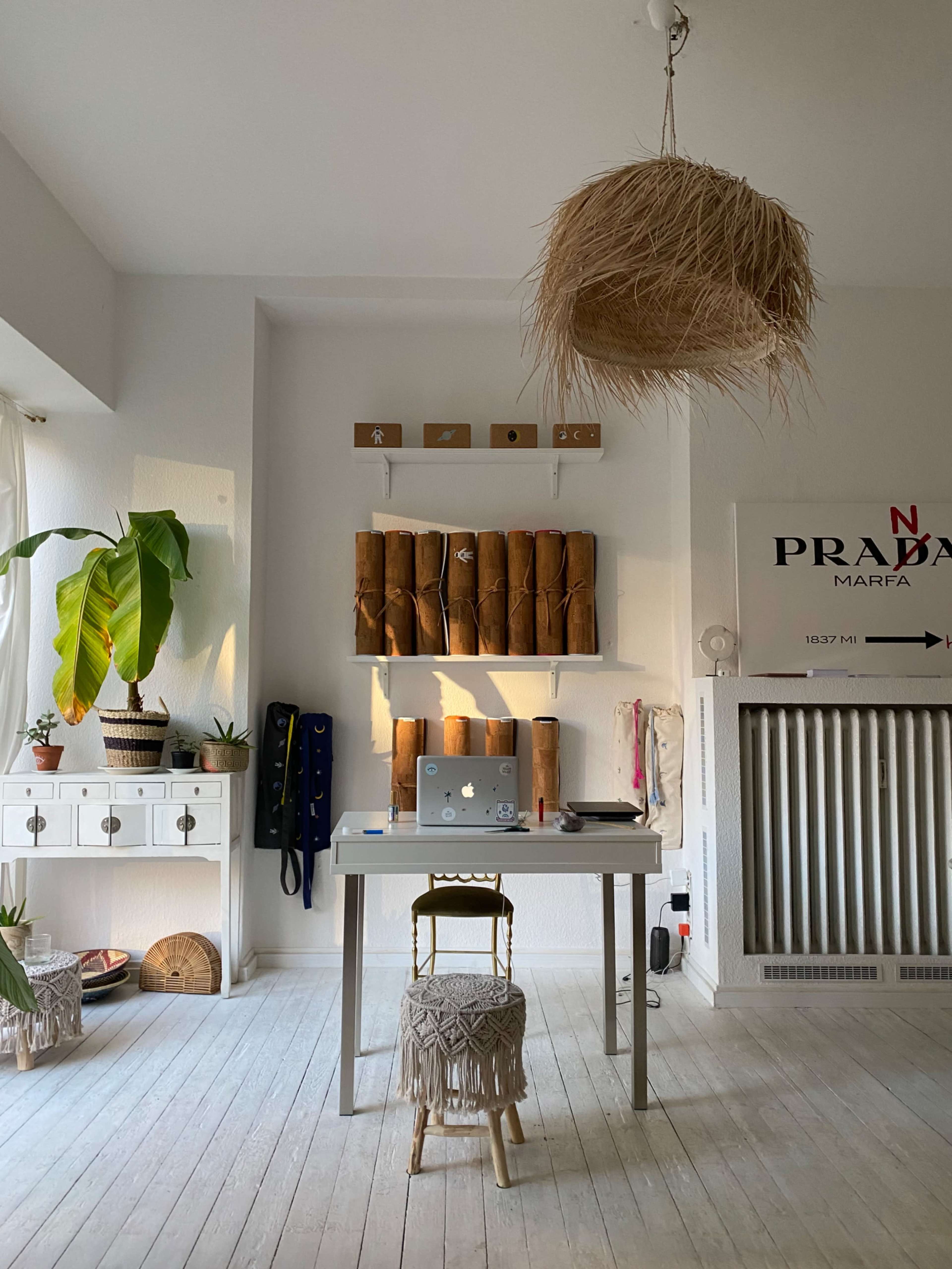 The image shows a minimalist workspace featuring a table with a laptop, surrounded by rolls of paper or fabric on shelves, a potted plant, and a decorative hanging light.