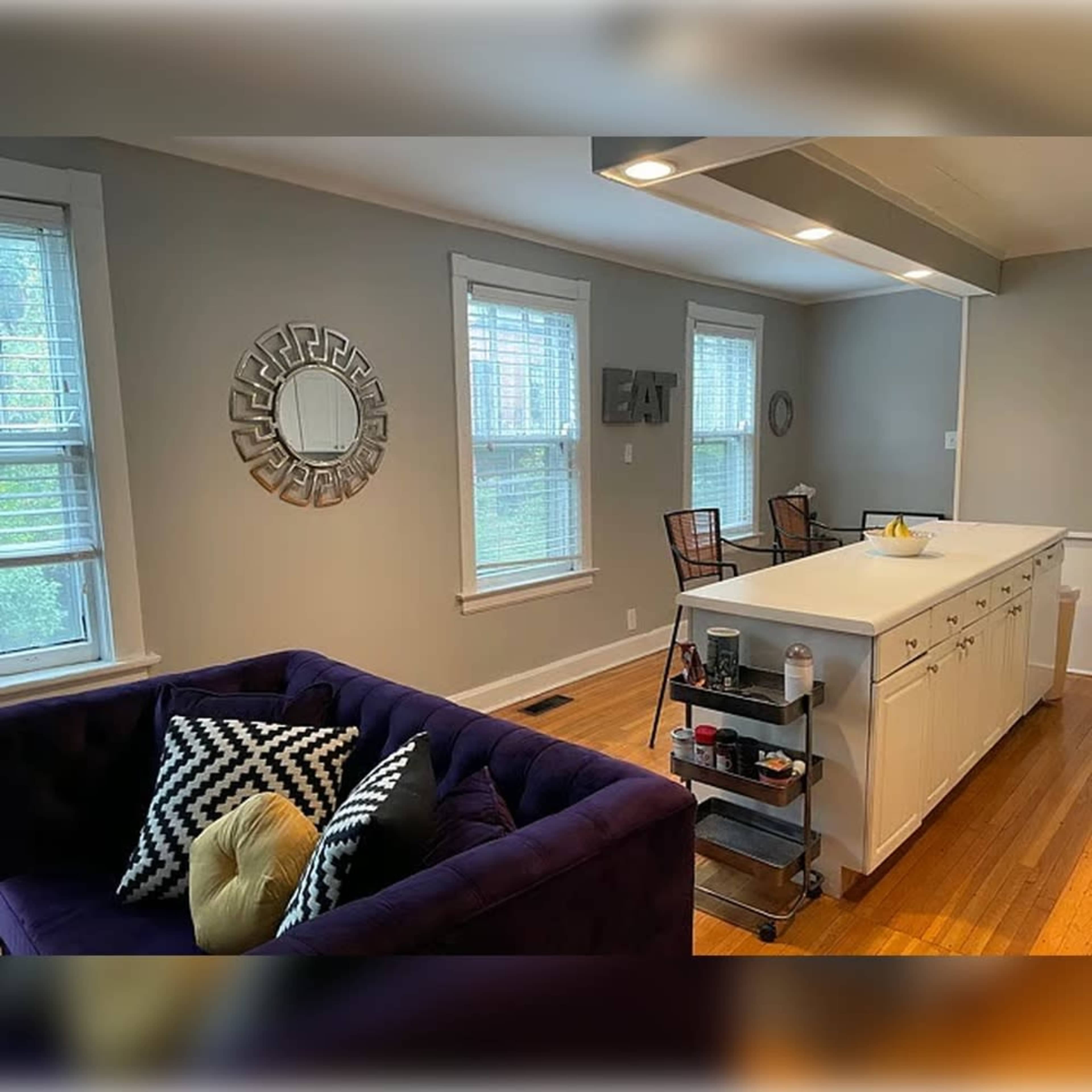 The image shows a modern living space featuring a purple couch, a kitchen island, and two windows allowing natural light to enter.