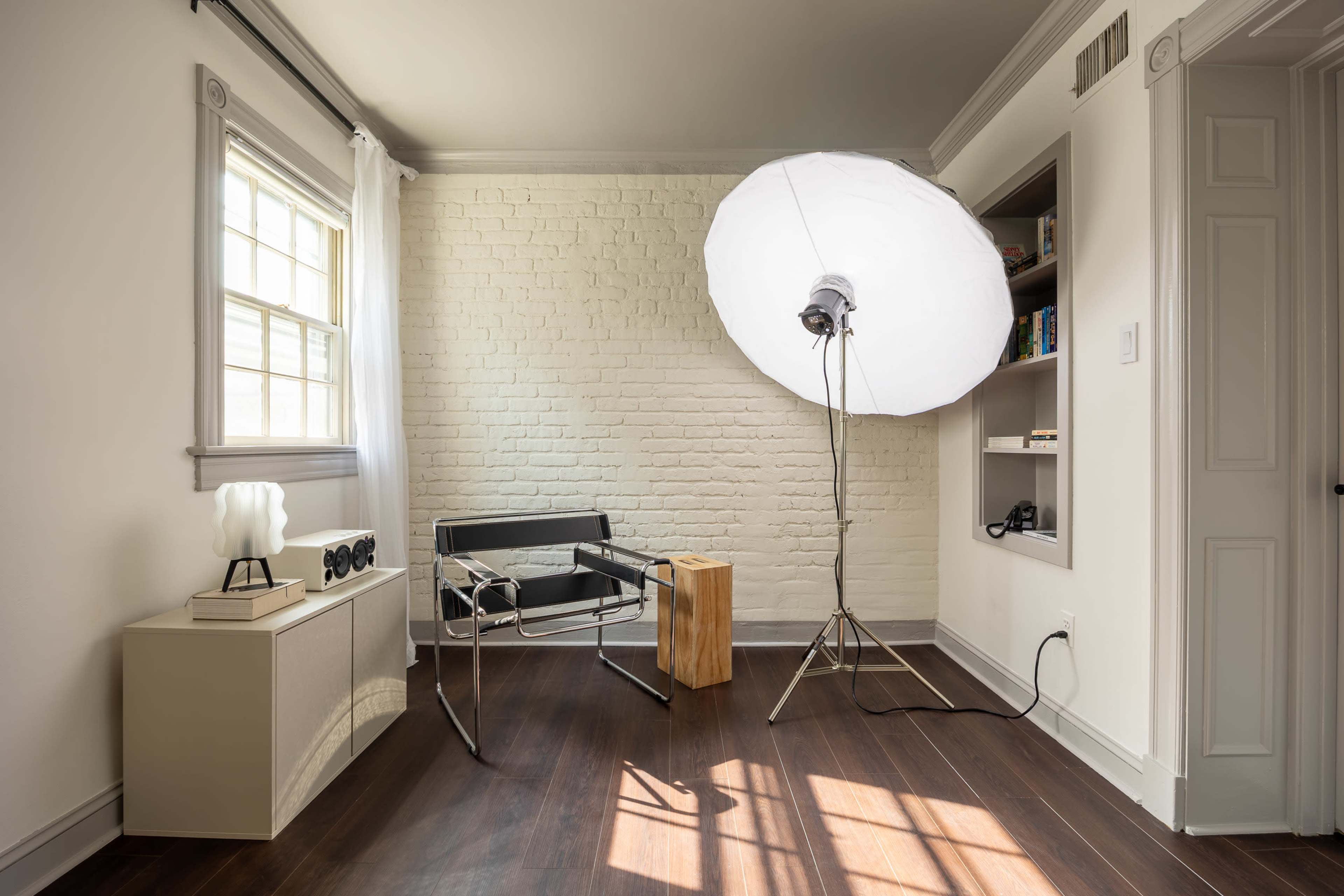 The image shows a well-lit room with a white brick wall, featuring a modern chair, a side table, and a large softbox light on a stand.