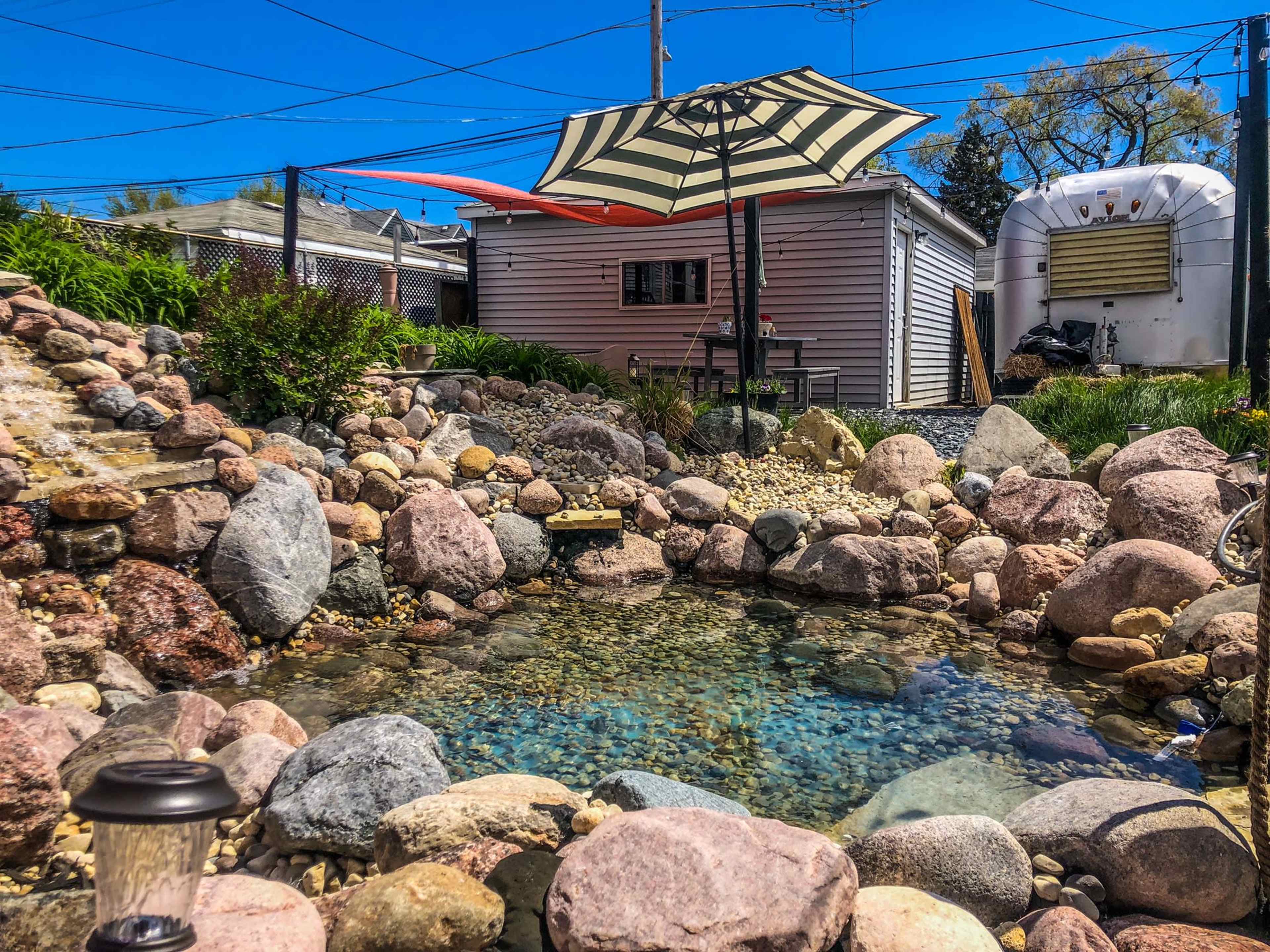 A landscaped backyard with a small stone pond surrounded by rocks and an umbrella over a seating area next to a shed.