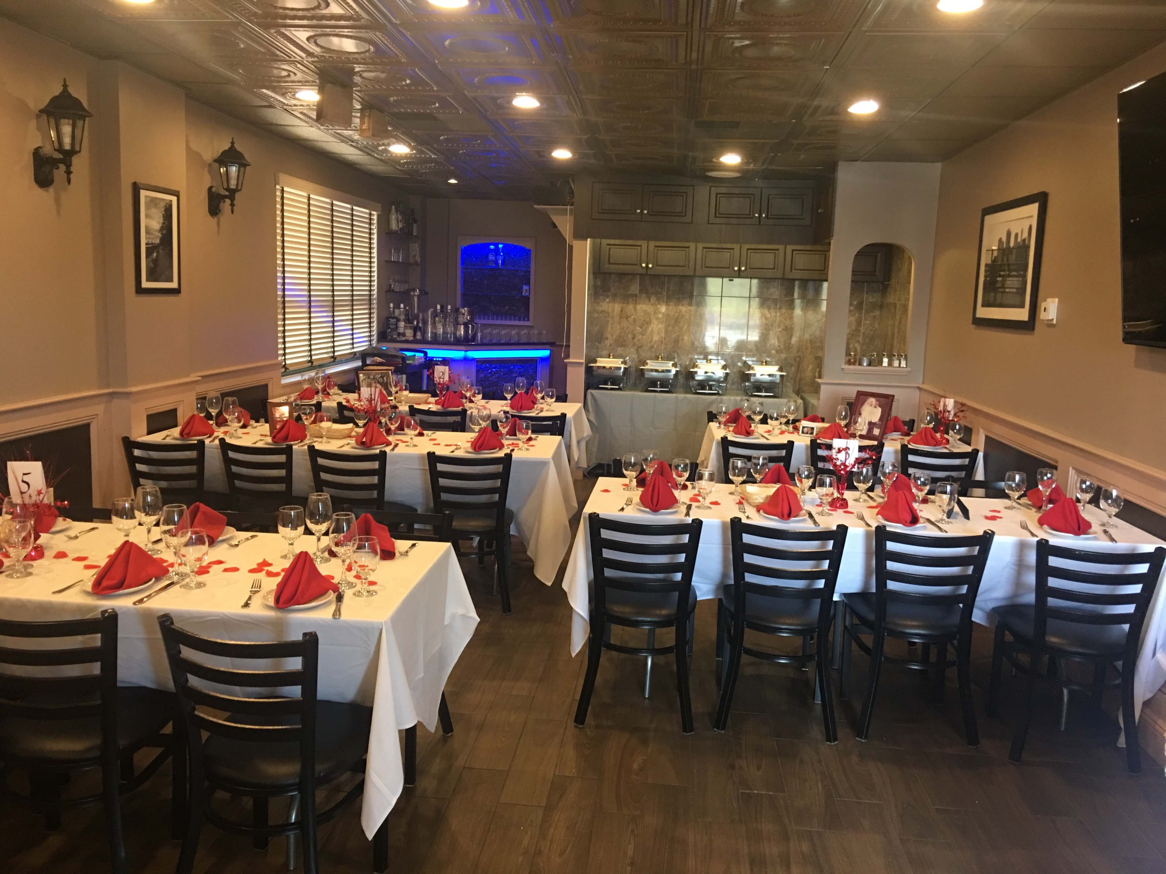 The image shows a dining area set up for an event, featuring several tables covered with white tablecloths, red napkins, and glassware, with a buffet area in the background.