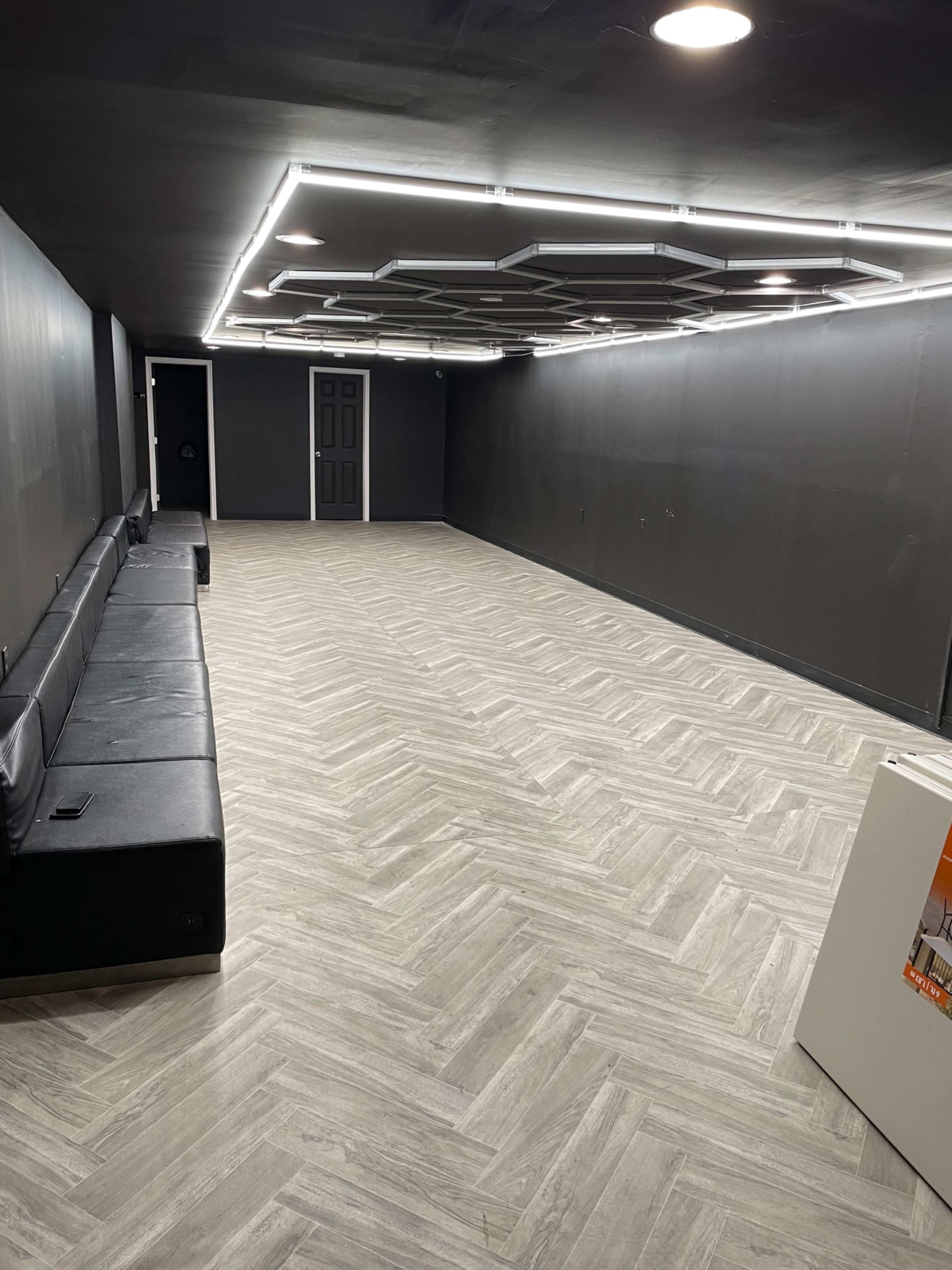 A spacious, empty room with a black wall, a herringbone-patterned floor, and a long black couch along one side.