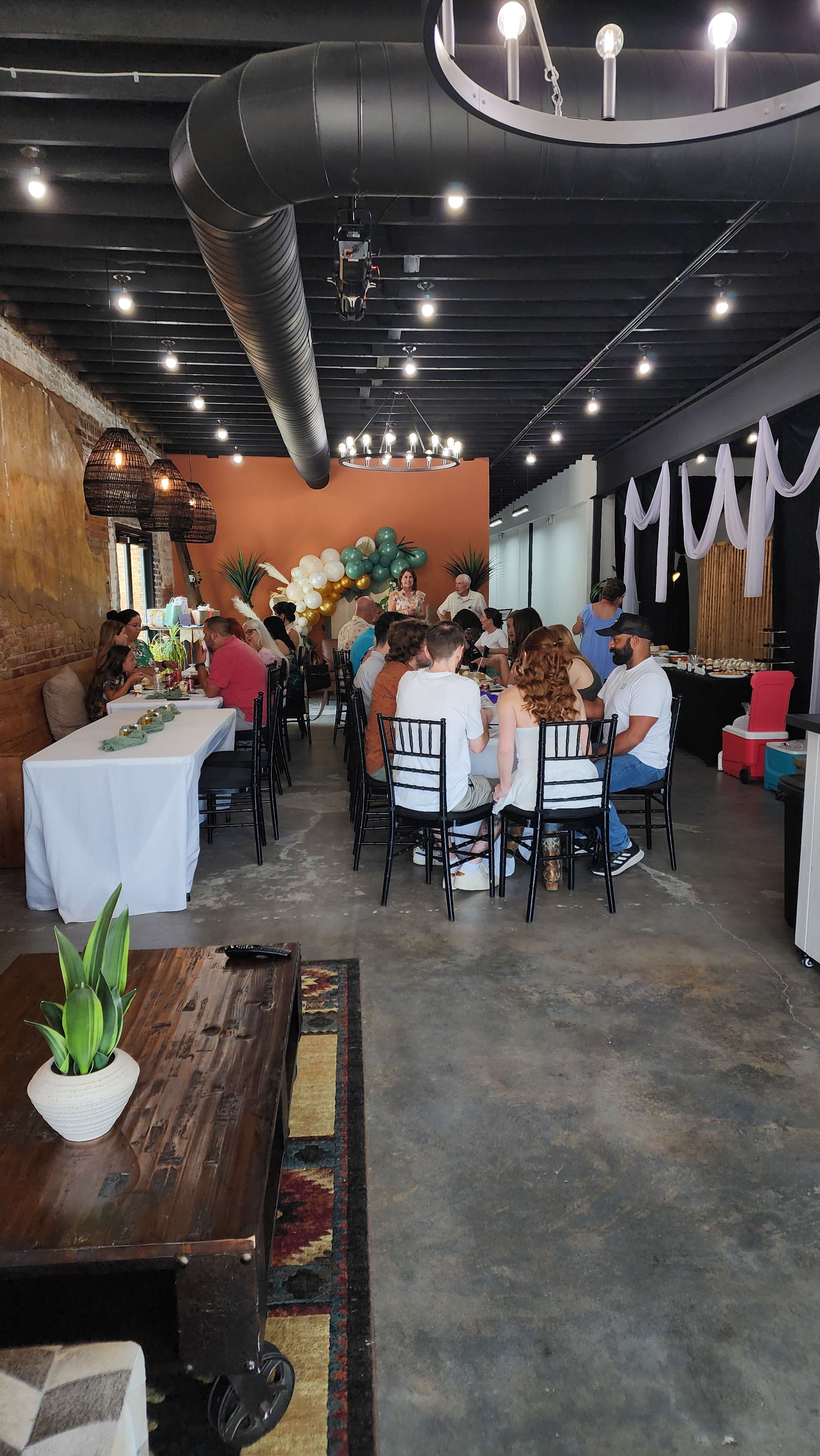 A dining area is set up with guests seated at long tables, surrounded by decorative balloons and plants.