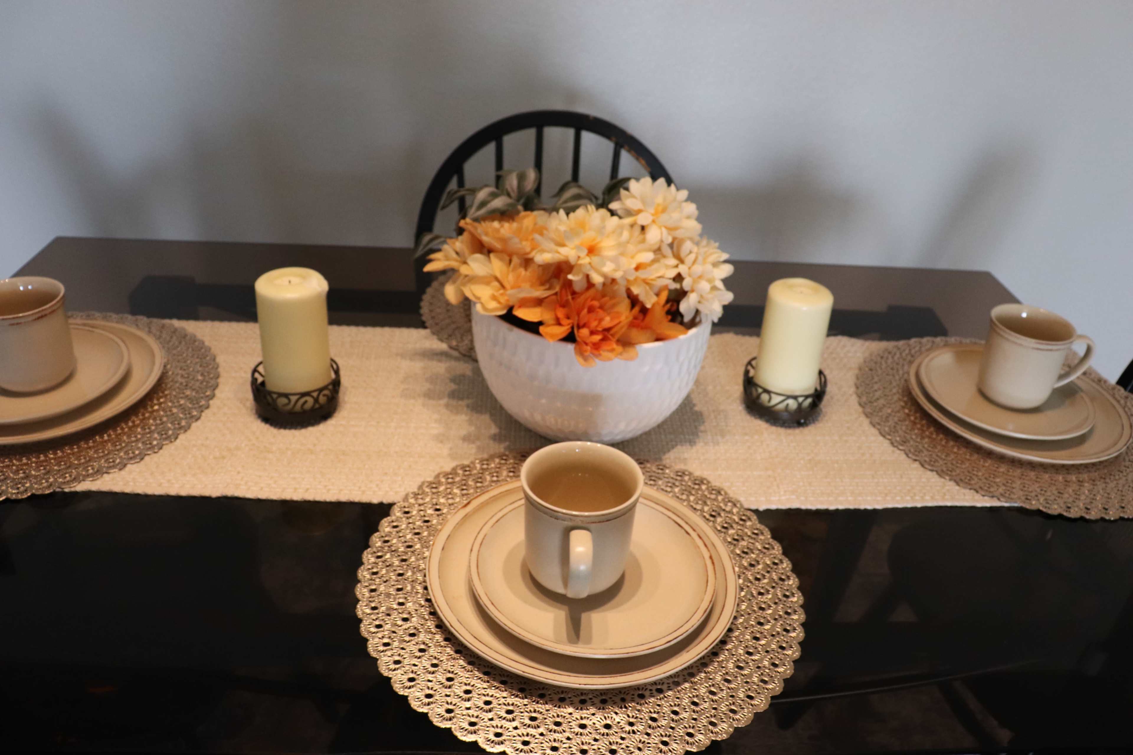 A dining table is set with four place settings, each featuring a plate and cup, along with a decorative bowl of flowers and candles in the center.