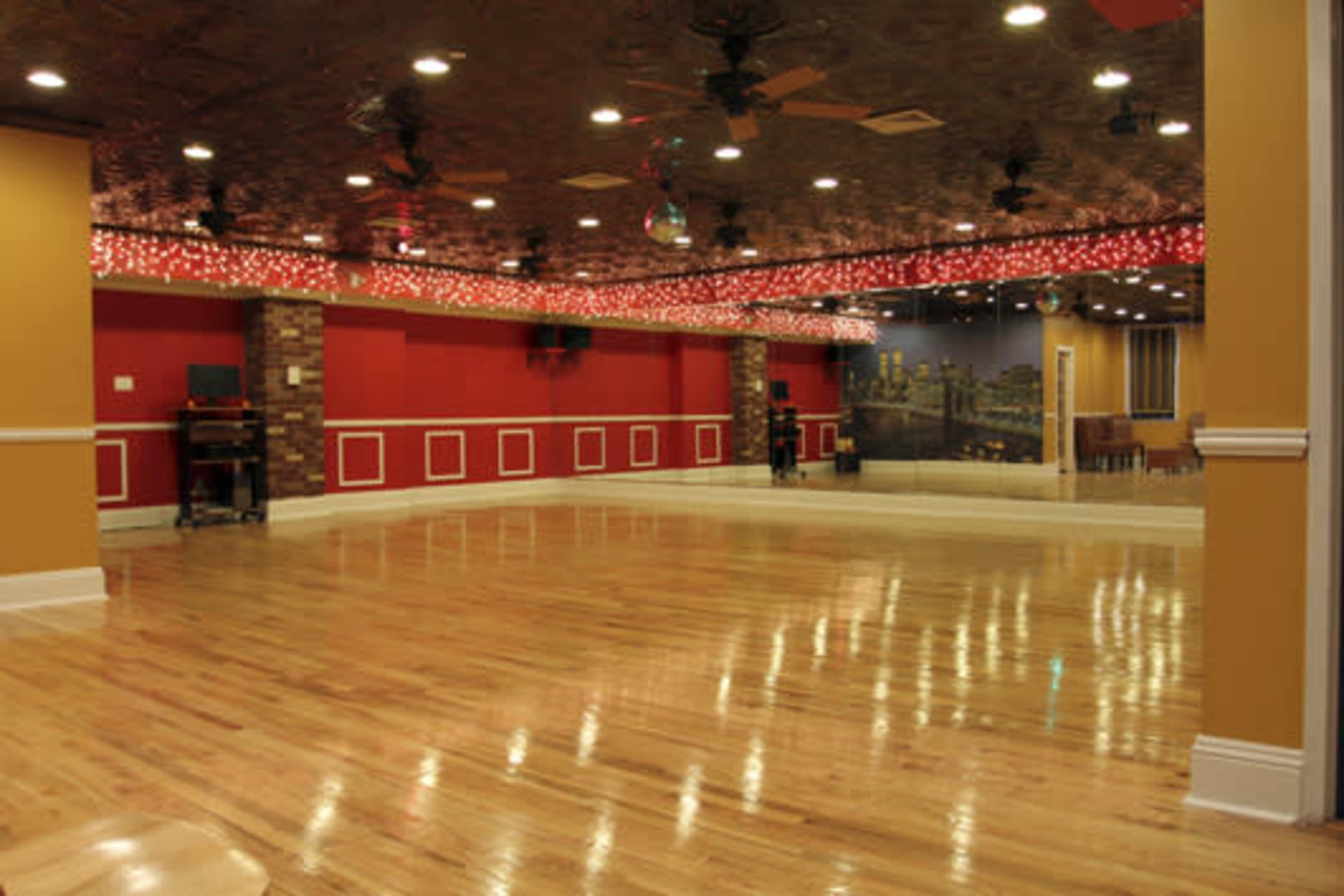 The image shows a spacious dance studio featuring wooden floors, red and yellow walls, and string lights along the ceiling.
