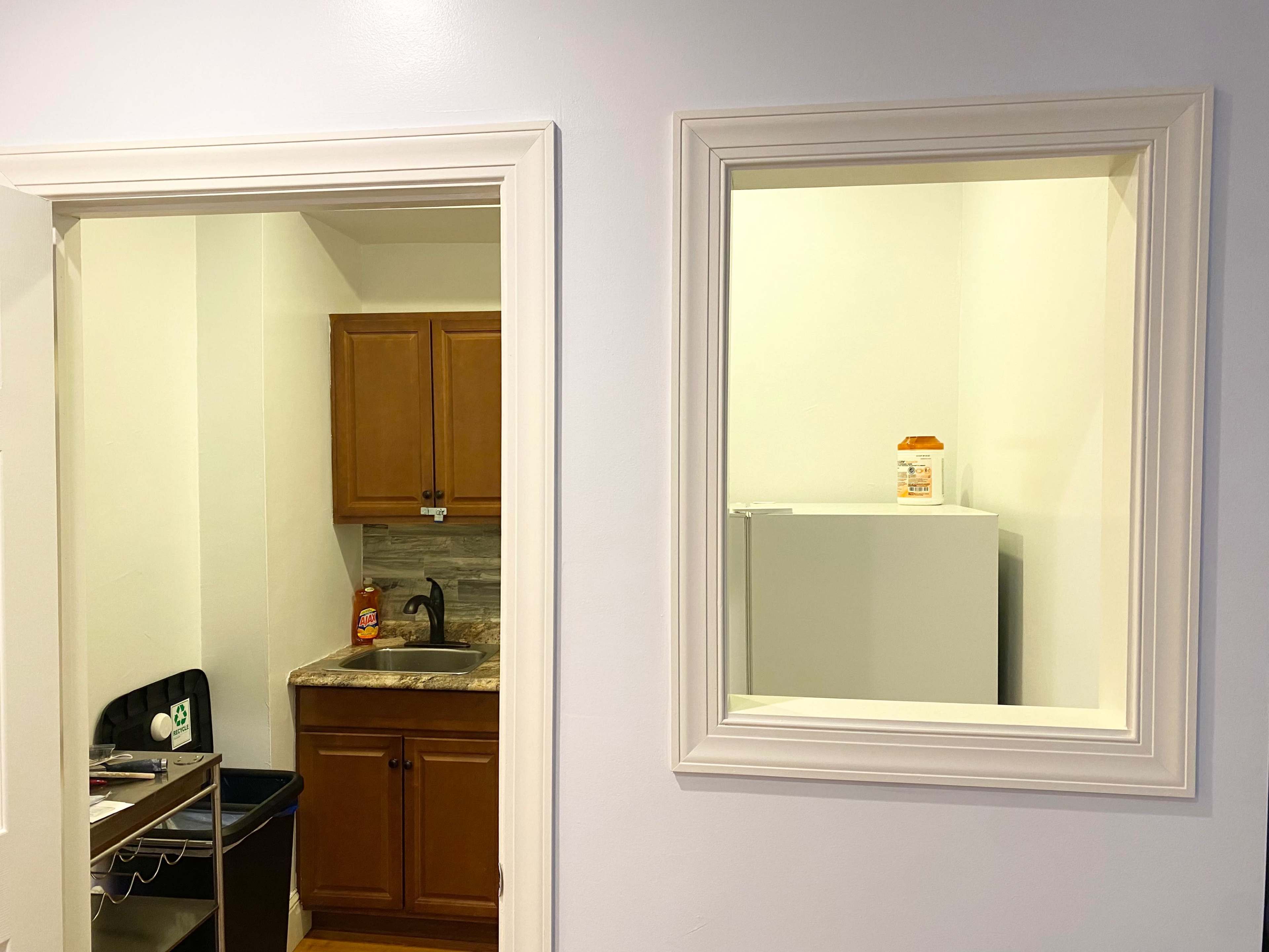 The image shows a kitchen area with wooden cabinets and a sink on one side, and a mirror-framed opening revealing a small room with a white refrigerator on the other side.