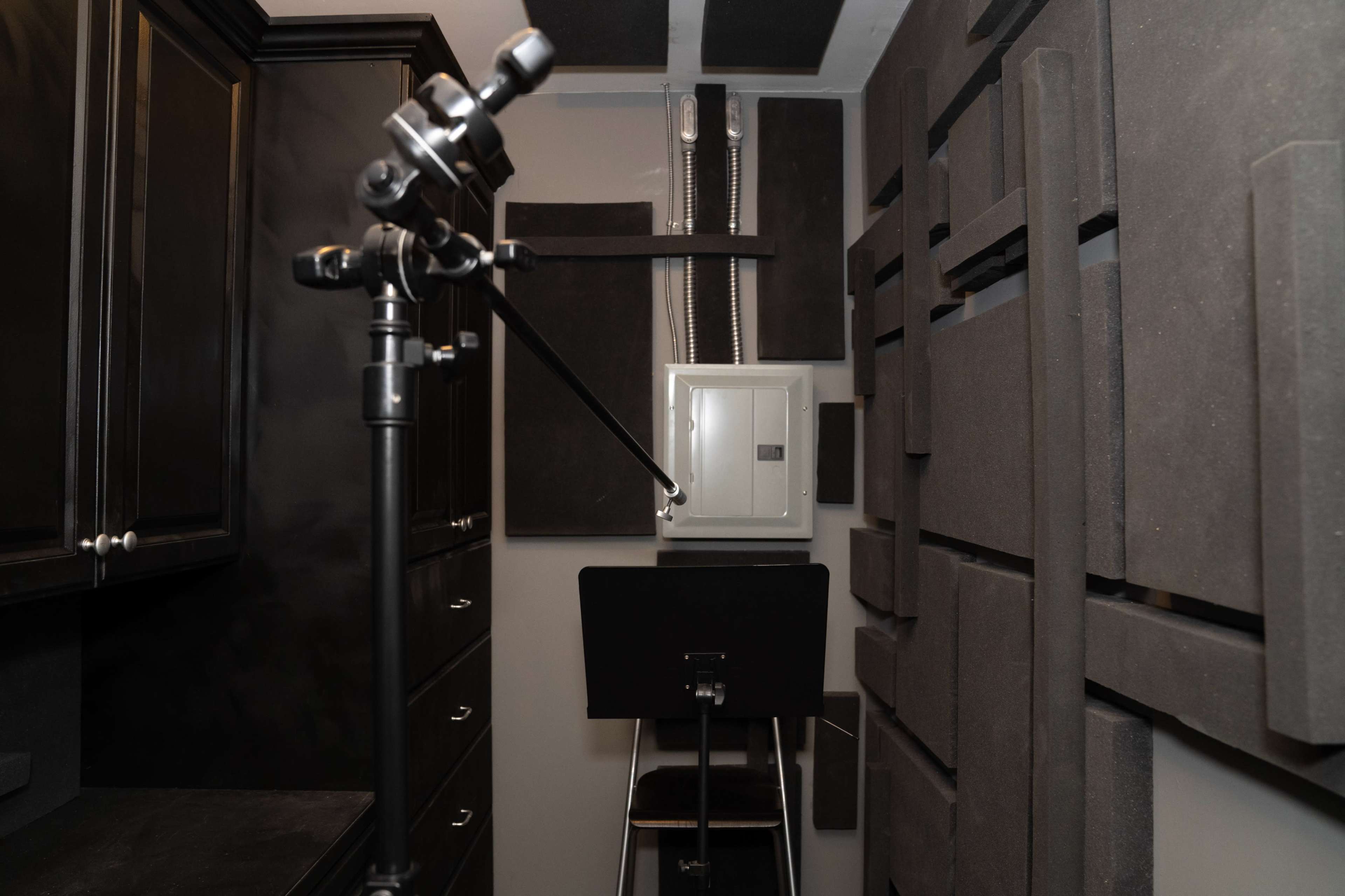 A small recording booth featuring soundproofing panels on the walls, a microphone stand, and a music stand in front of an electrical panel.