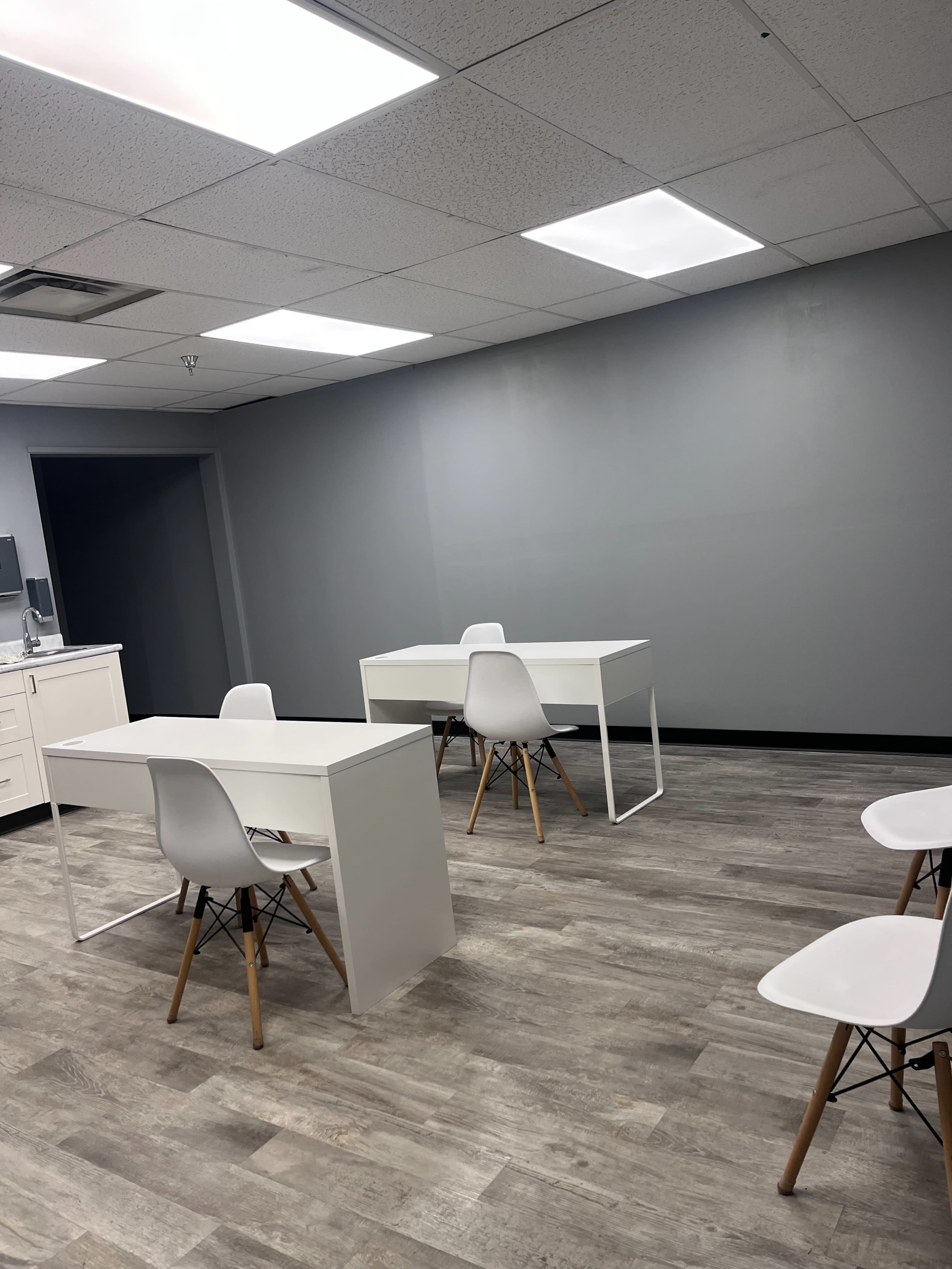 The image shows a minimally furnished office space with two white desks and matching chairs on a wood-patterned floor.
