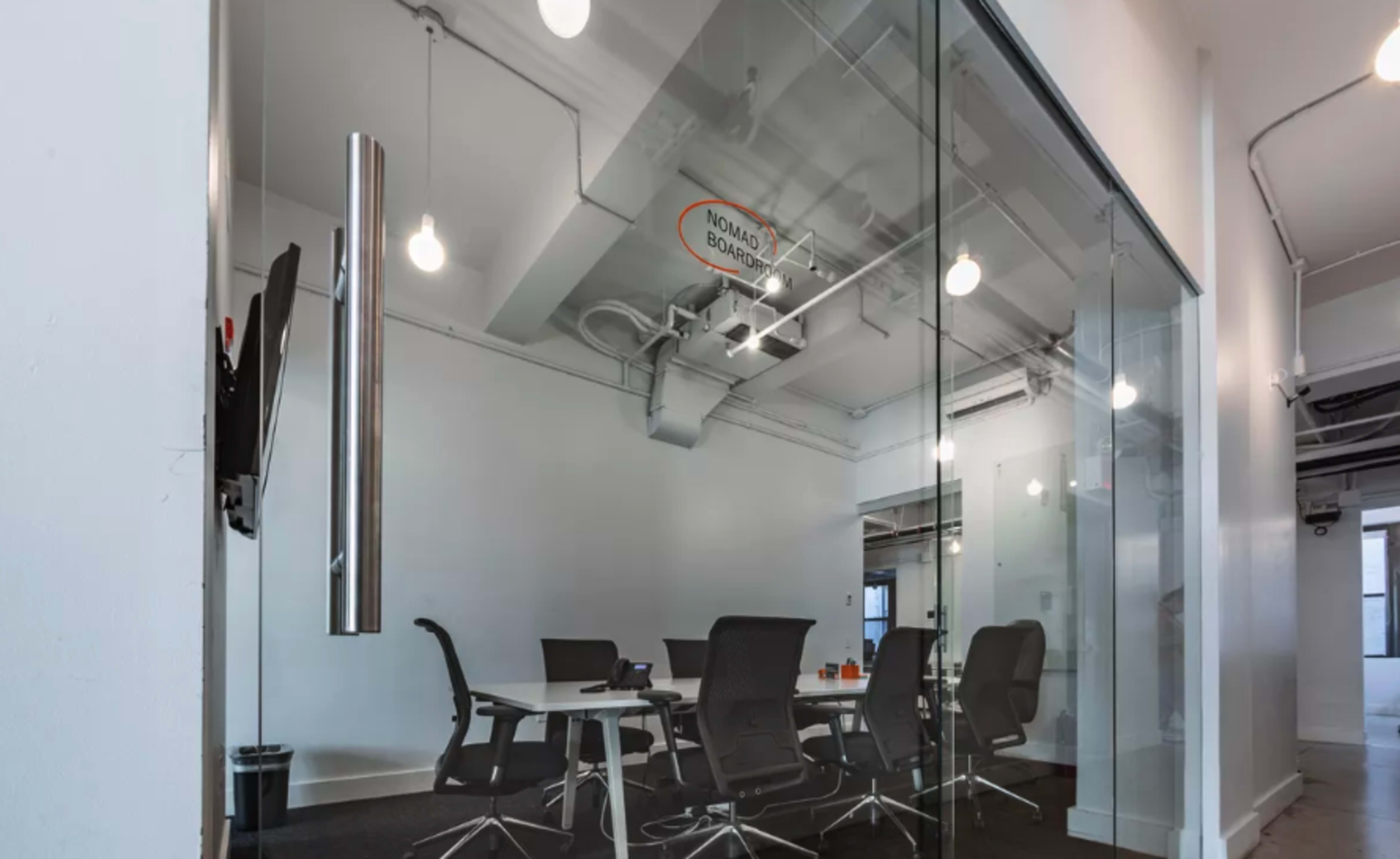 A modern conference room features a glass wall, a round table, and ergonomic chairs under bright overhead lighting.
