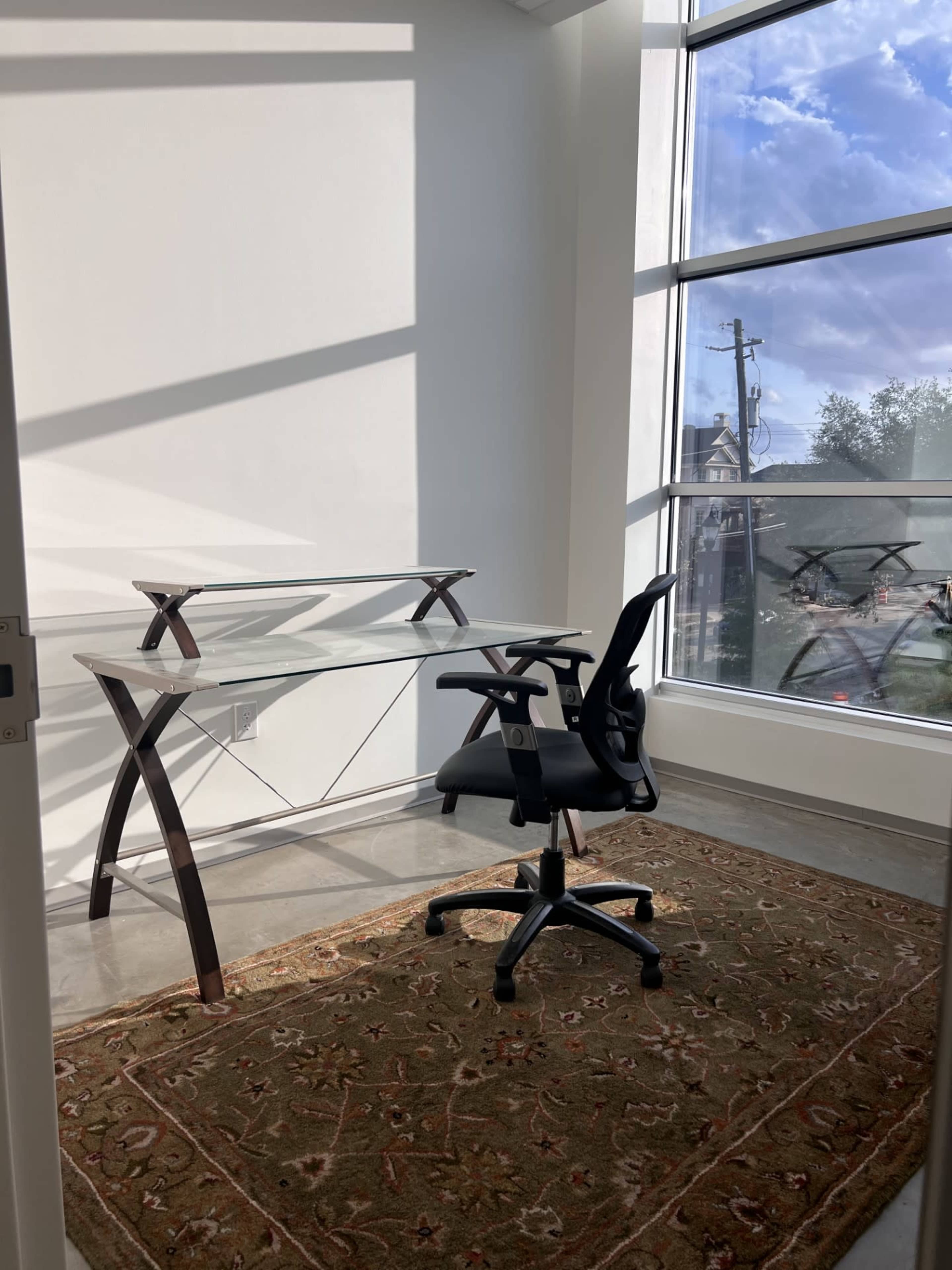 A modern glass desk with an ergonomic black chair is positioned near a large window, illuminated by natural light.