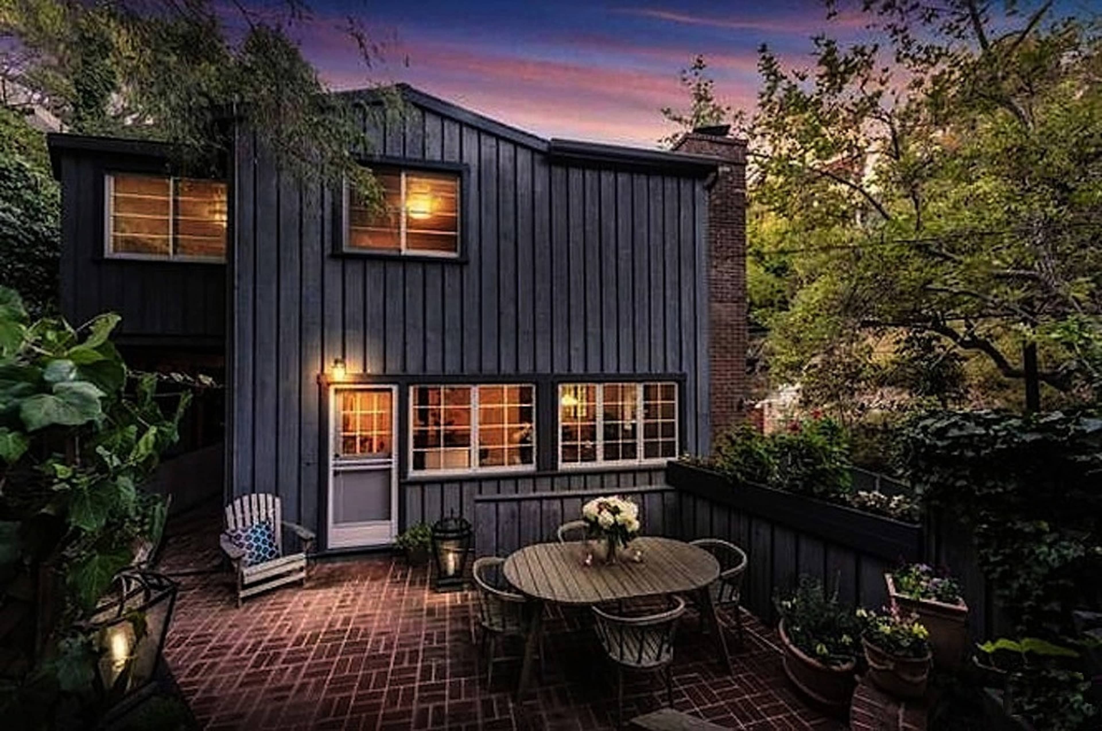 A two-story wooden house with a dark exterior features a patio area with a round table and chairs, surrounded by greenery at dusk.
