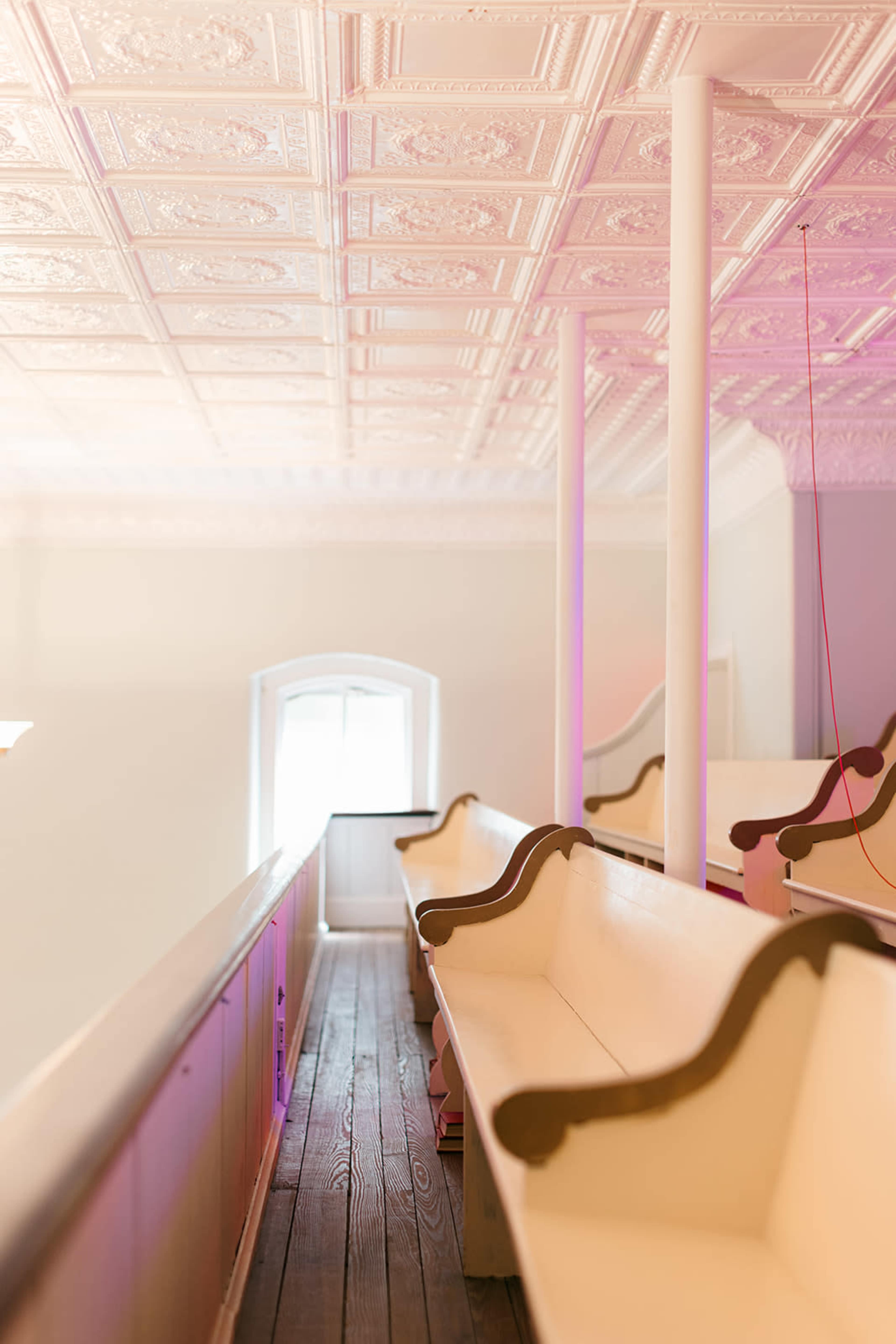 The image shows a well-lit, empty interior space featuring rows of wooden benches against a paneled wall and a decorative ceiling.