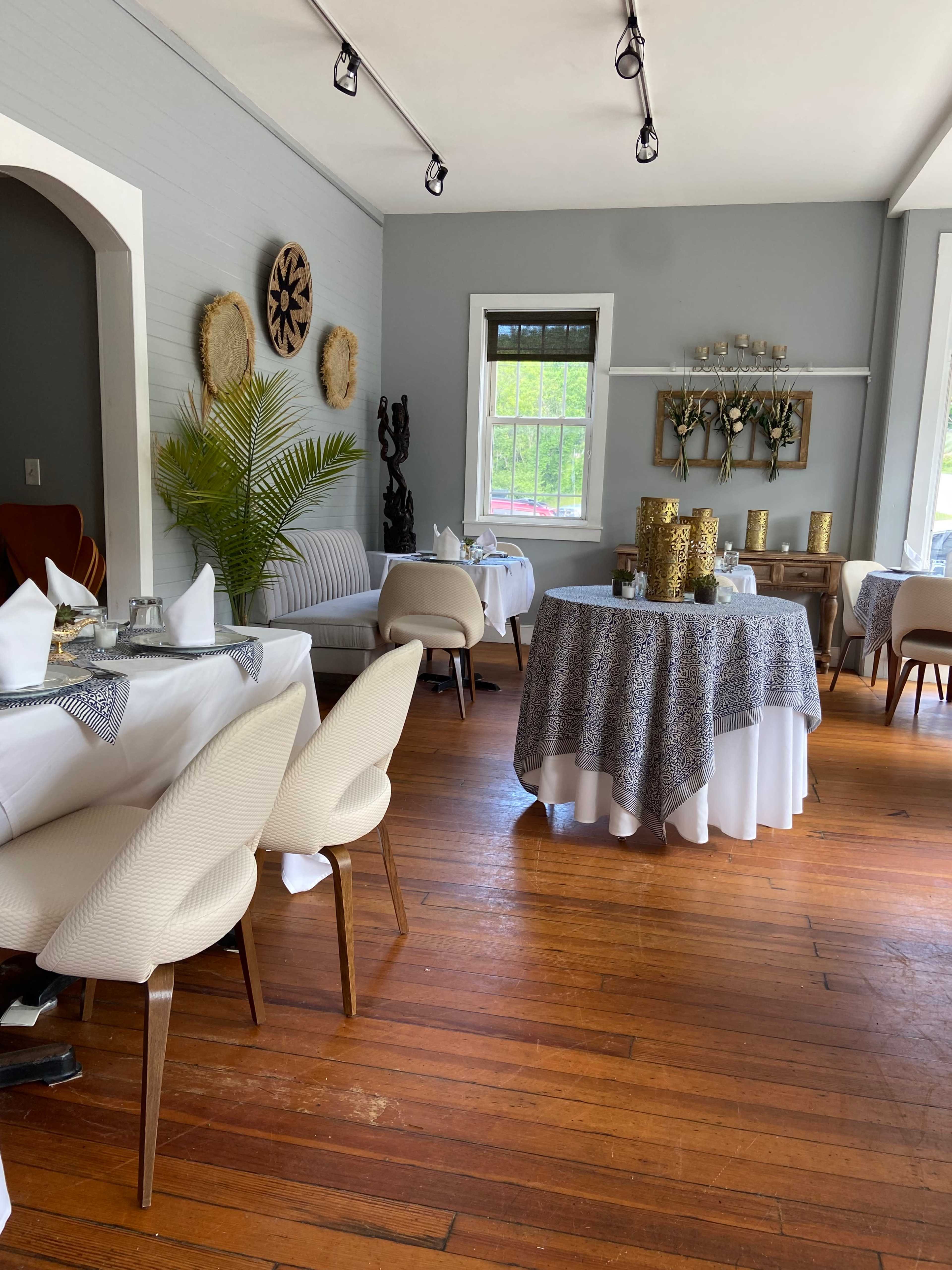 A dining room with wooden floors, round and rectangular tables set with white tablecloths, and light-colored chairs arranged around them.