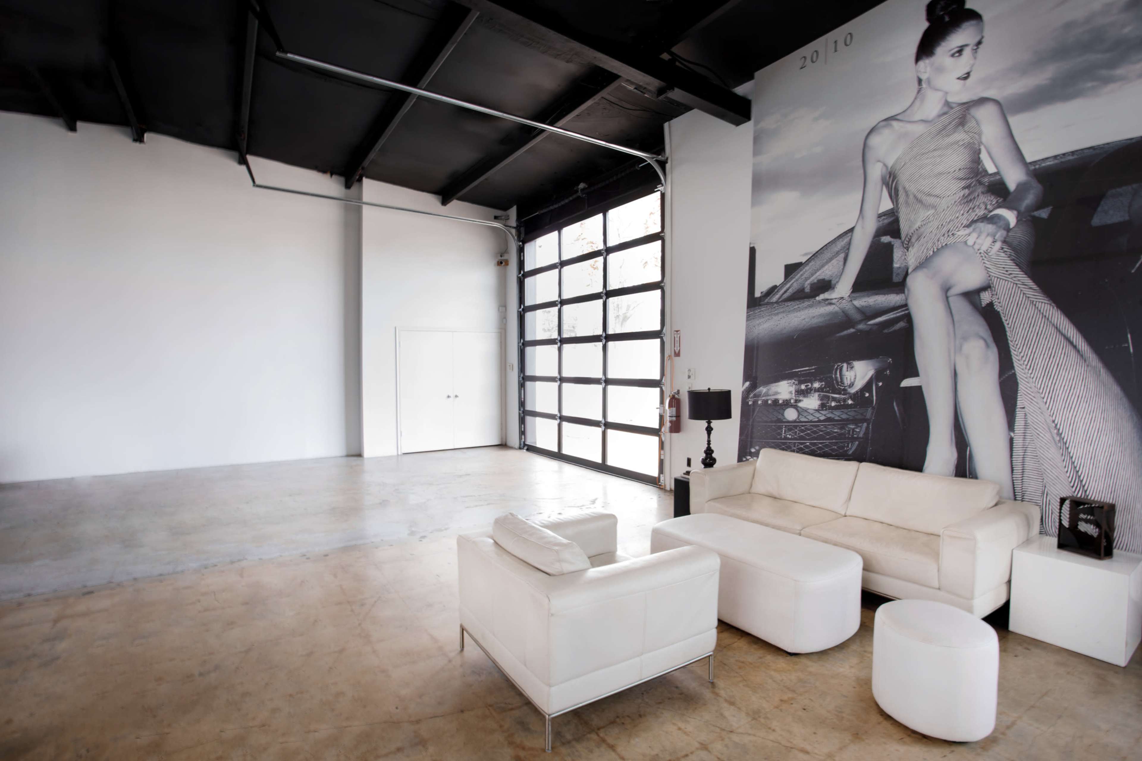 A minimalist interior with a white sofa set and large glass doors featuring a large wall mural of a woman beside a car.