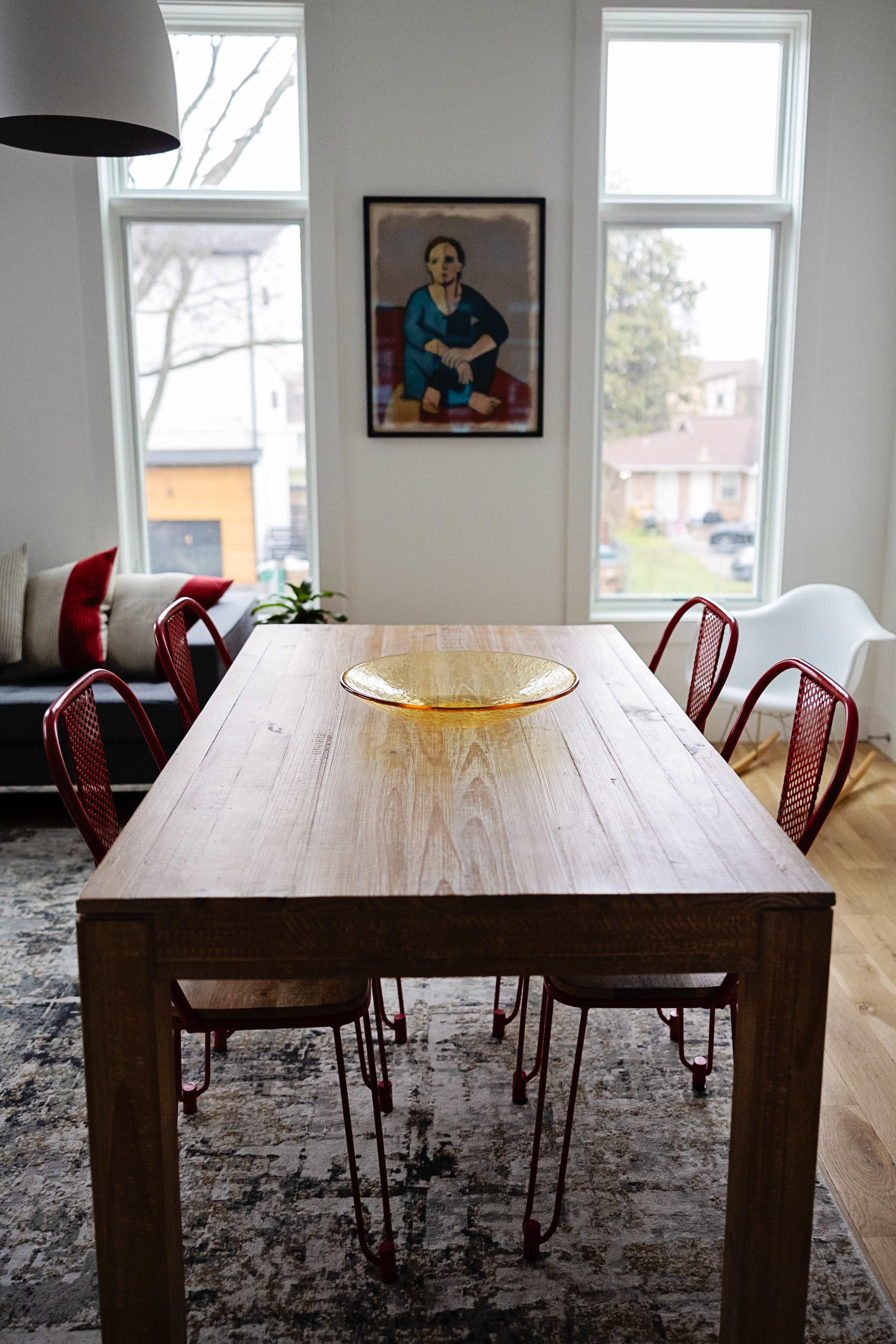 A wooden dining table is set in a room with large windows, surrounded by red chairs, and features a decorative bowl in the center.