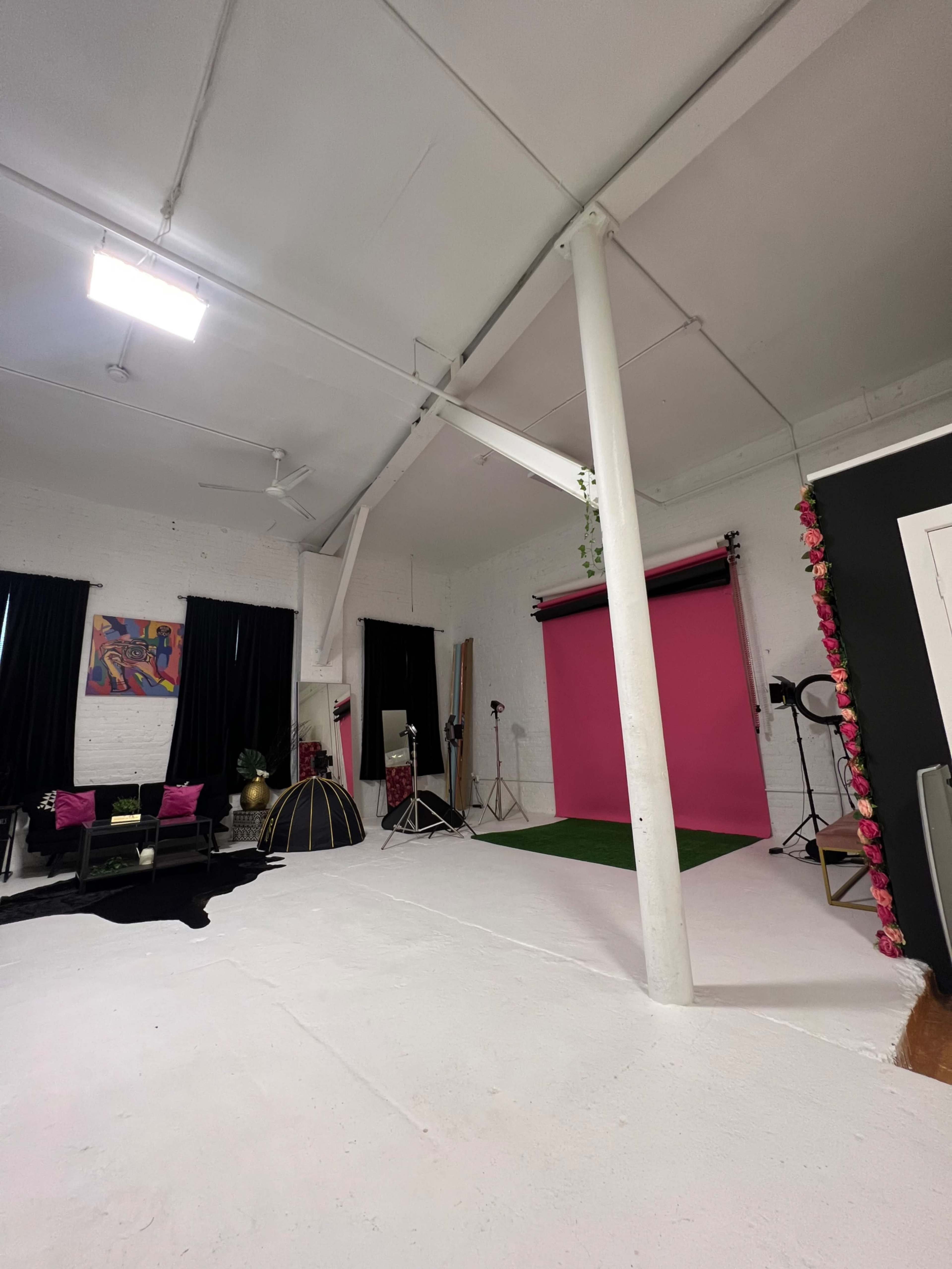 A photography studio with a pink backdrop, green carpet, and various lighting equipment set up, alongside black curtains and furniture.