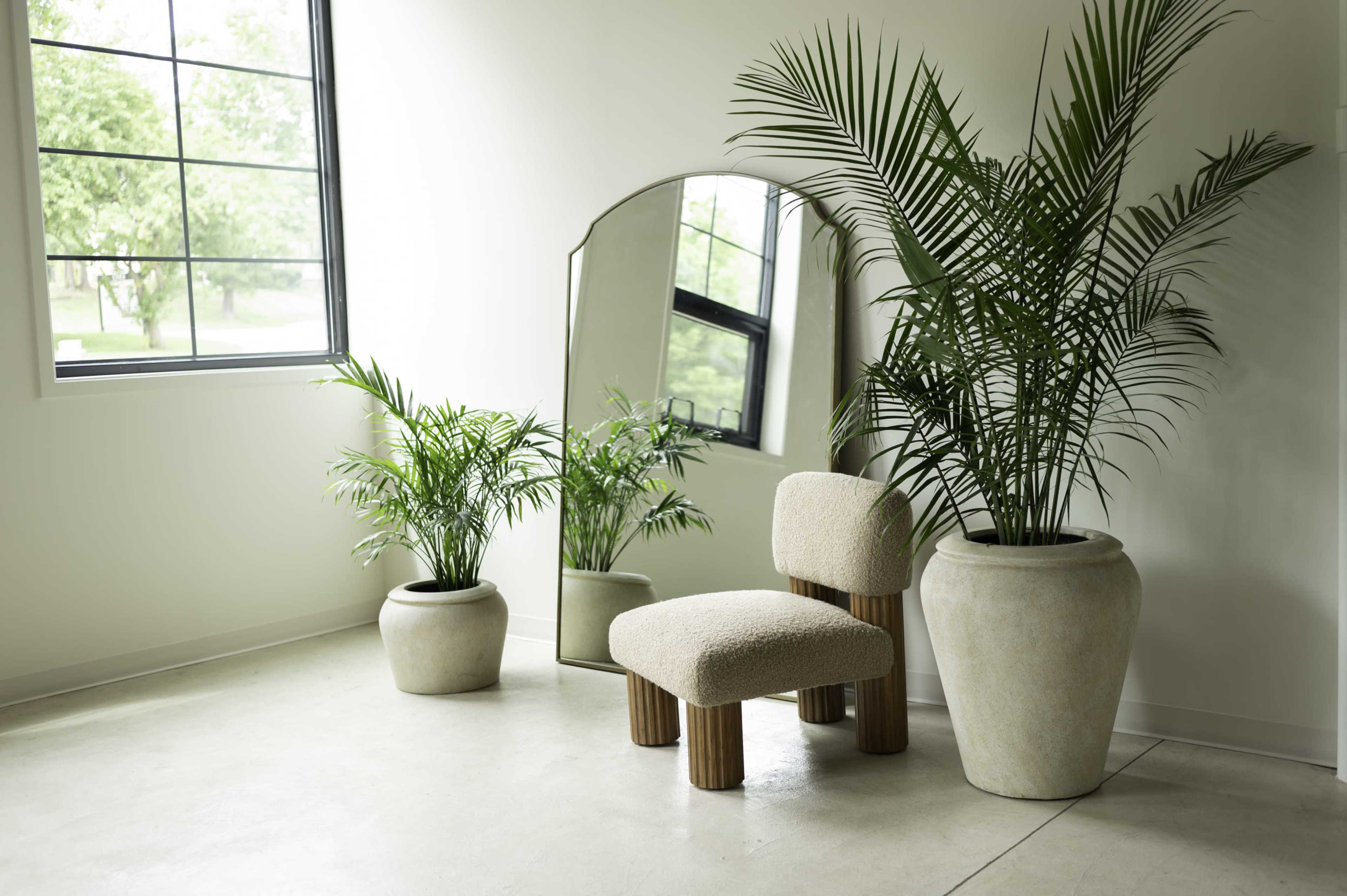 A corner of a bright room features a large mirror, a plush chair, and two potted plants.