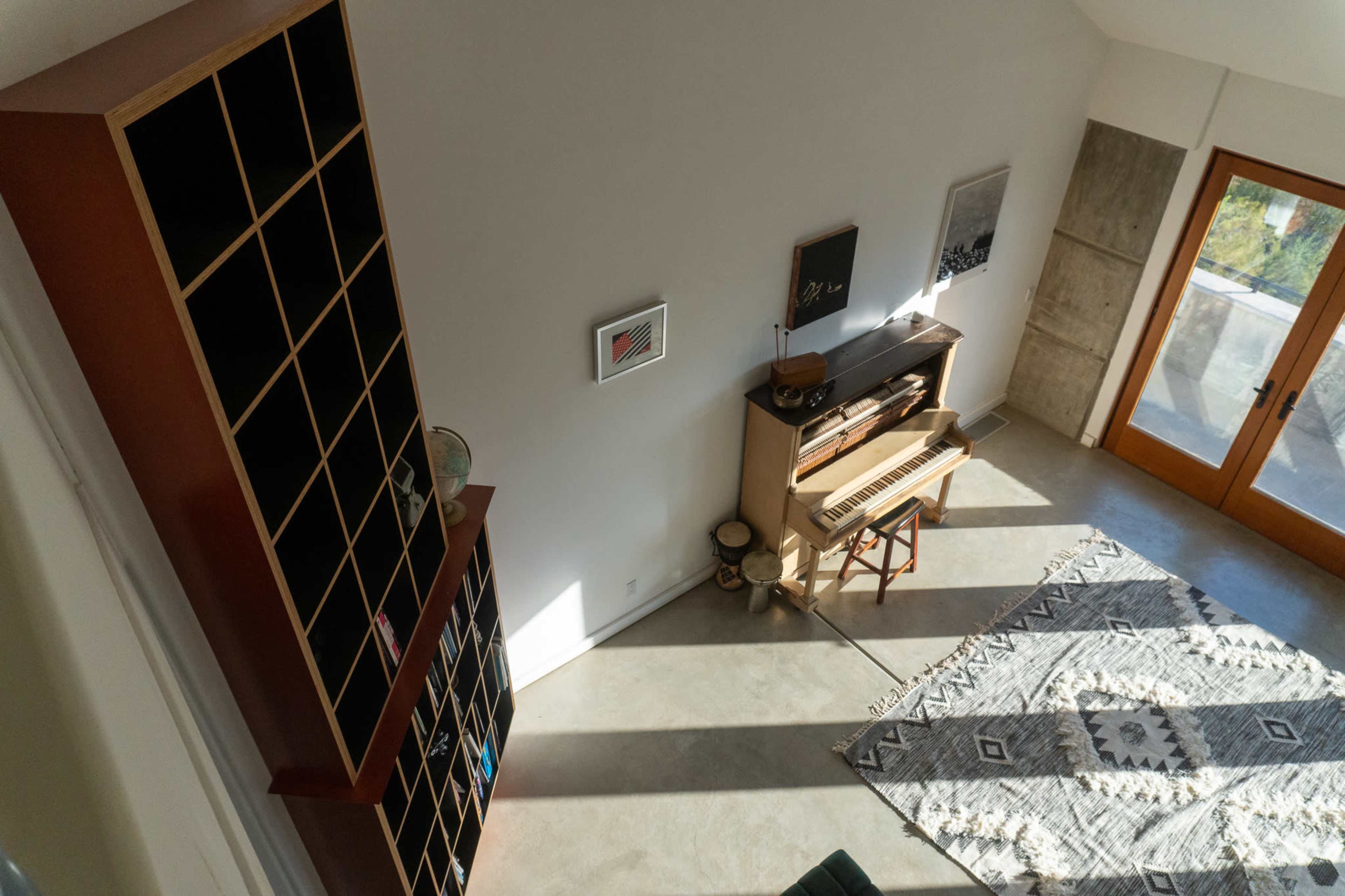 An overhead view of a modern room featuring a piano, a bookshelf with cubbyholes, and large windows allowing natural light to enter.