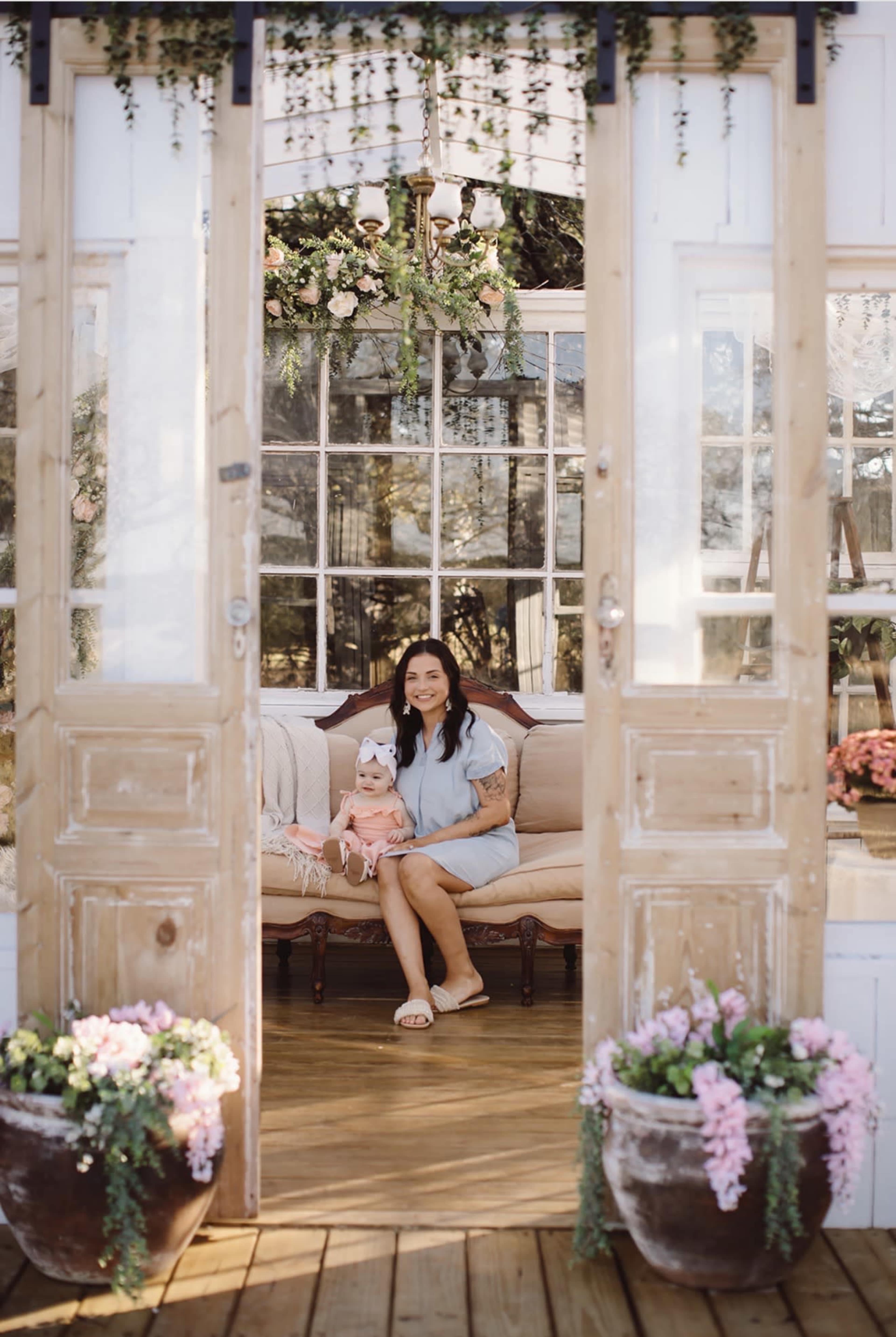 A woman holding a baby sits on a couch inside a glass-paneled room adorned with flowers and greenery.