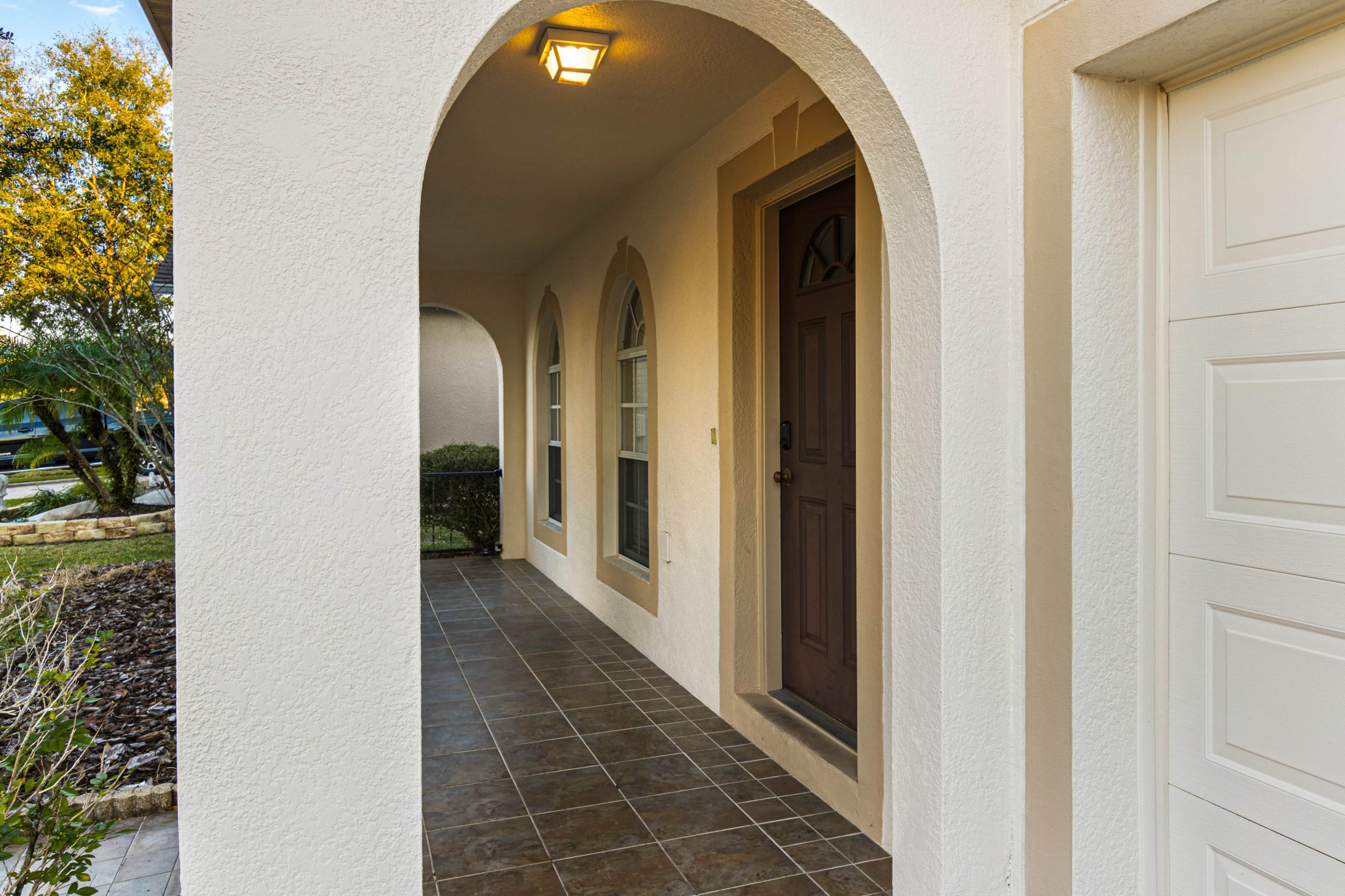 A covered entryway with a brown door and arched windows, leading to a porch with tiled flooring.