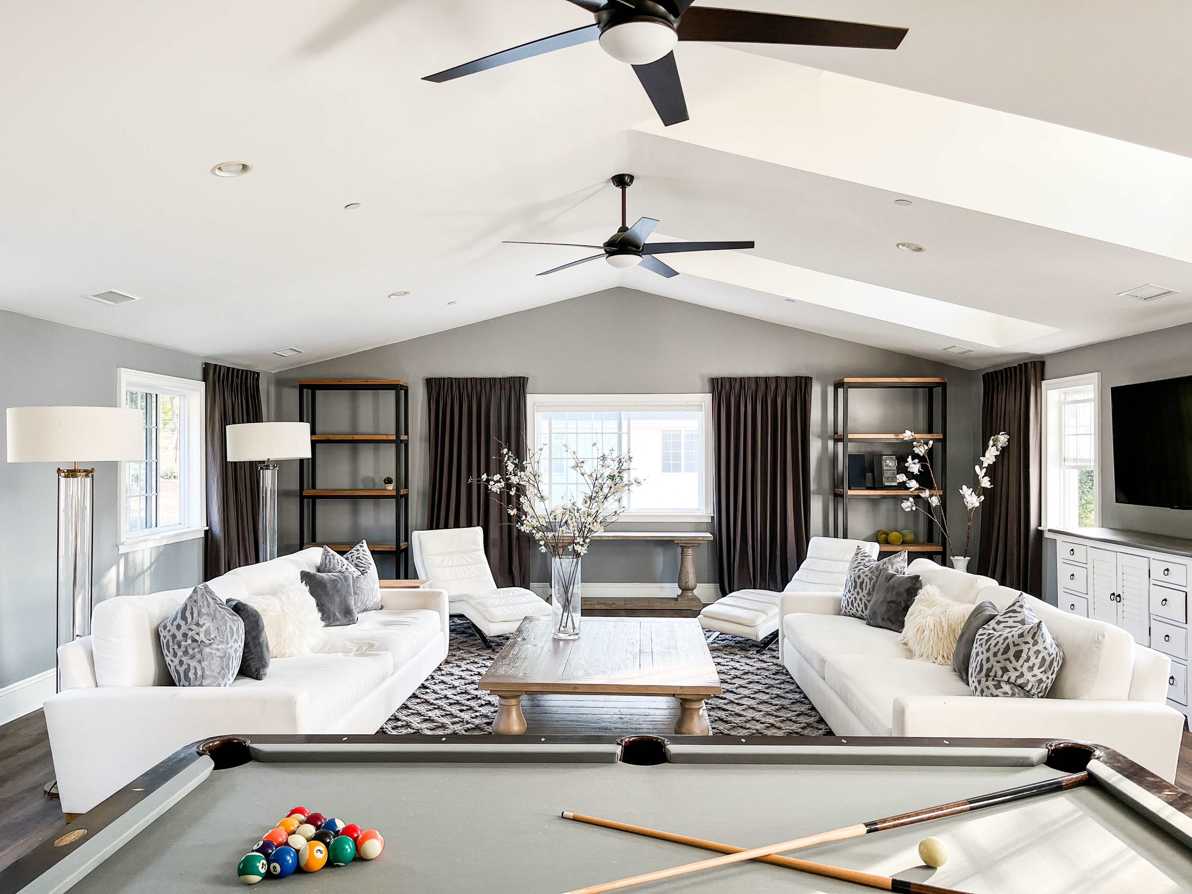 A spacious, modern living room features two white sofas facing a wooden coffee table, with large windows framed by dark curtains and decorative shelves.