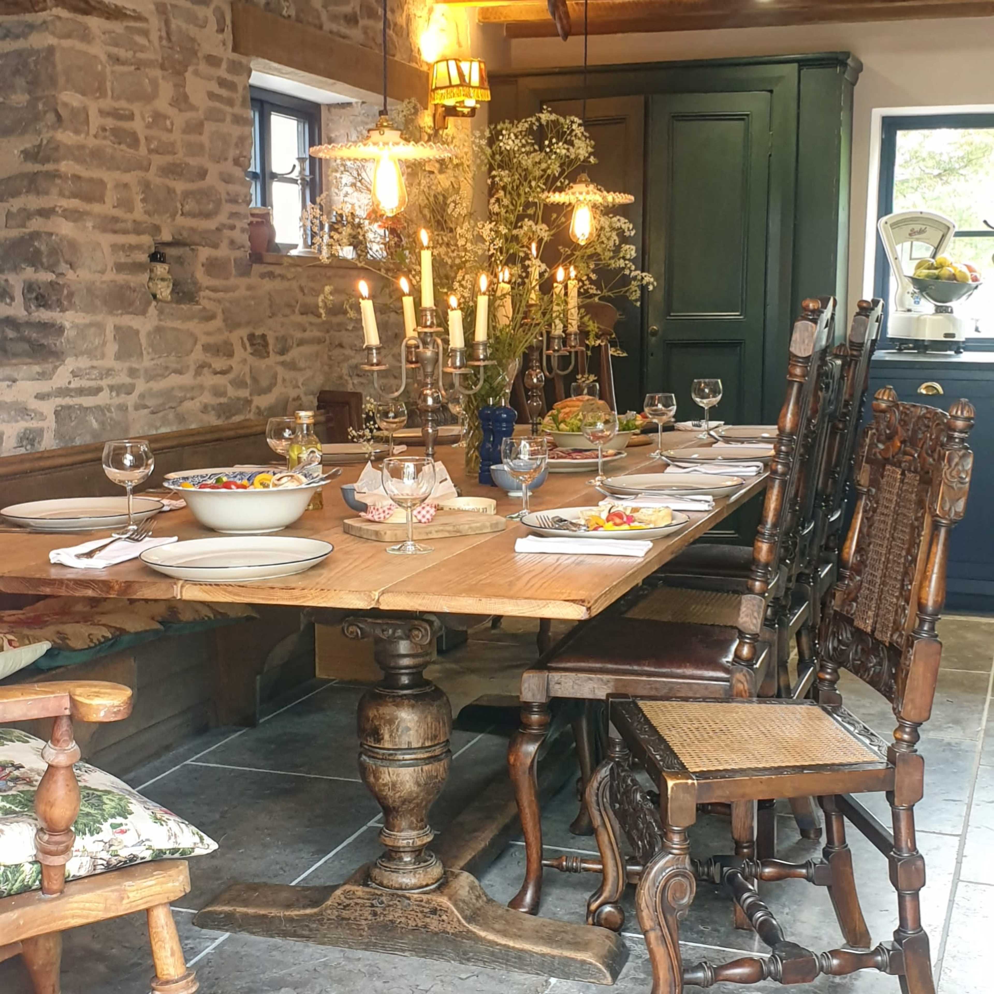A rustic dining room with a large wooden table set for a meal, surrounded by wooden chairs and illuminated by pendant lights.