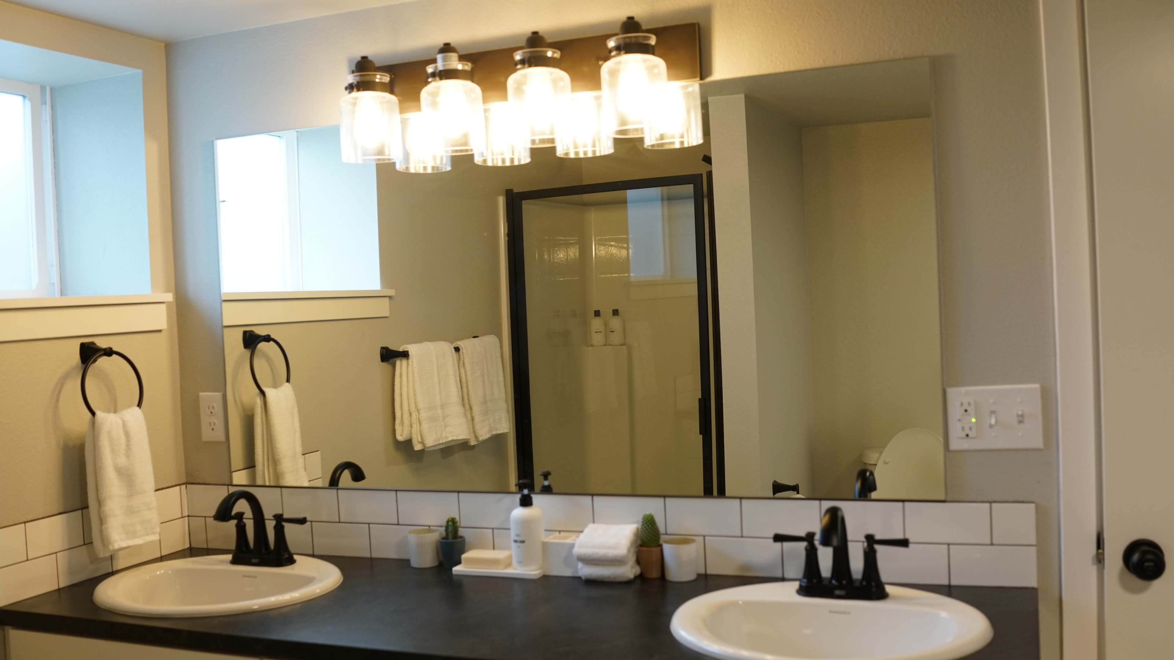 A bathroom features two sinks, a large mirror, and a shower enclosure, with pendant lights above and towels hanging on a rack.