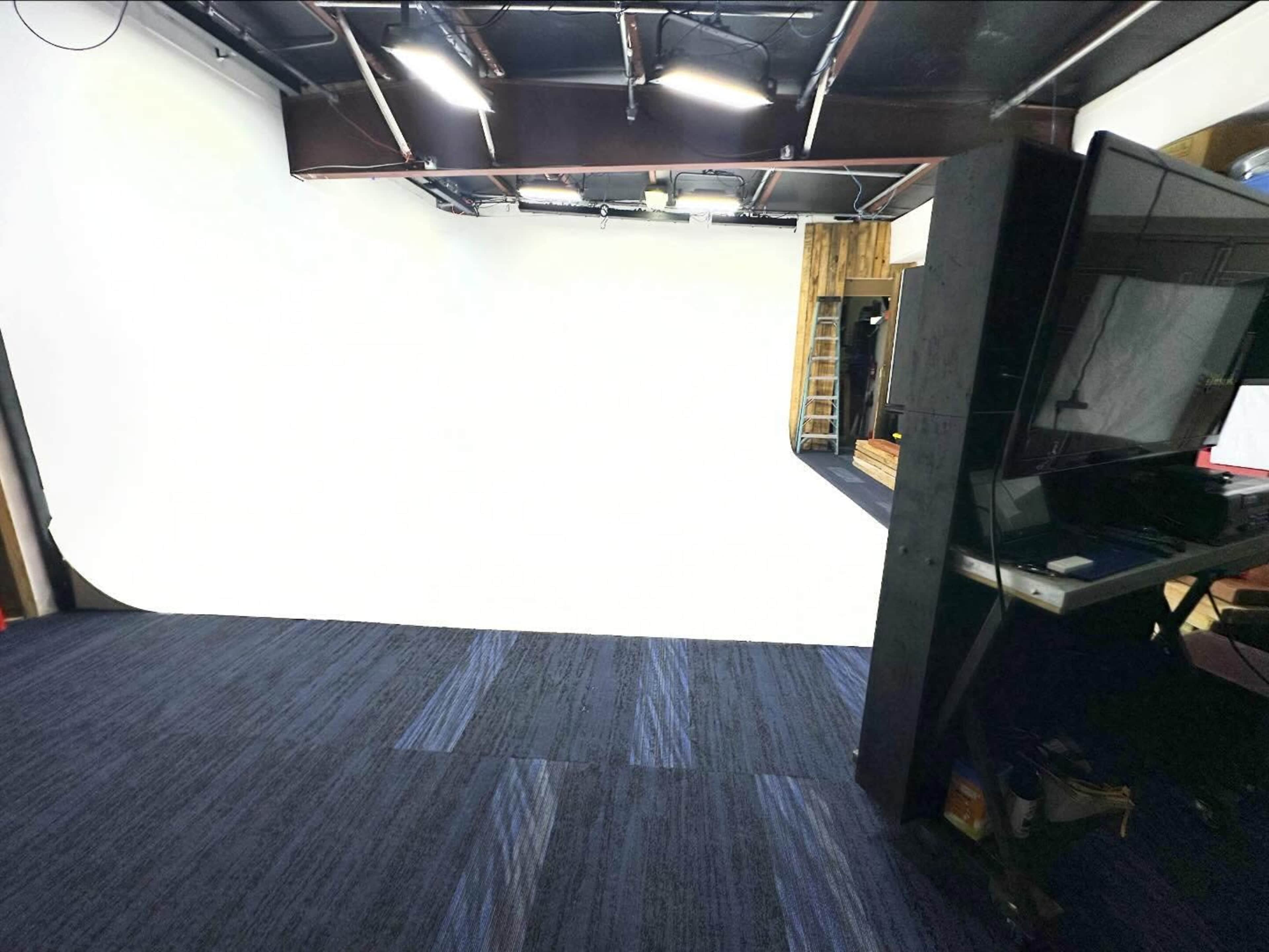 The image shows a photo studio with a large white backdrop, black overhead lighting, and a dark-colored floor.
