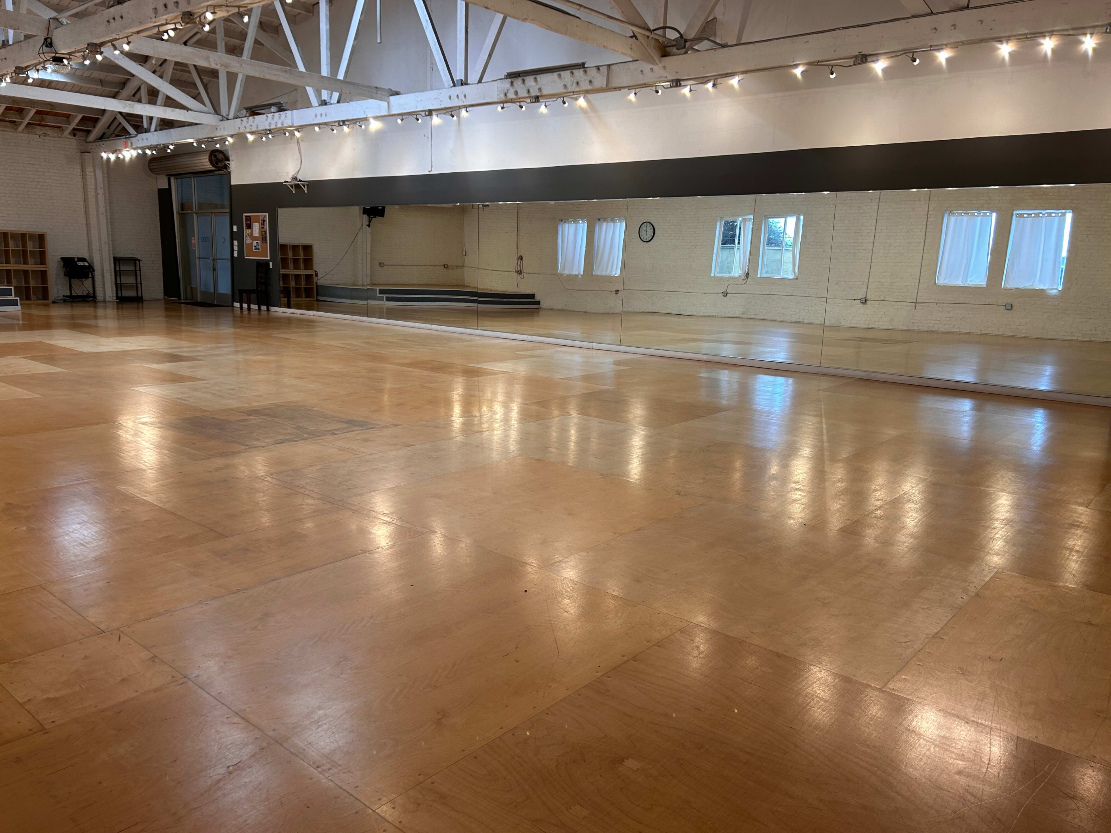 A spacious dance studio with a polished wooden floor, large mirrors, and bright lighting.