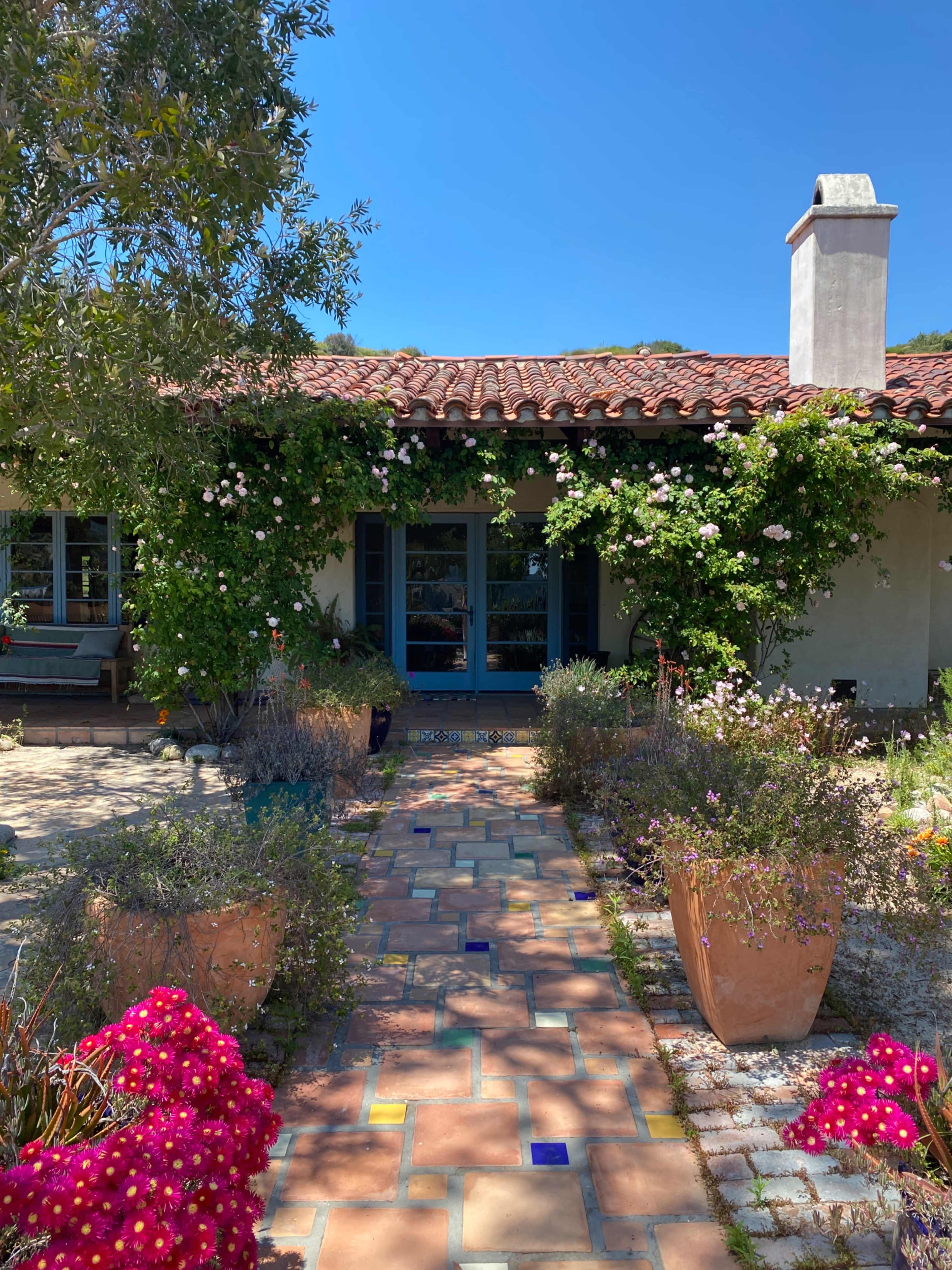 A path made of colorful tiles leads to a house adorned with climbing roses and surrounded by vibrant flowerpots.