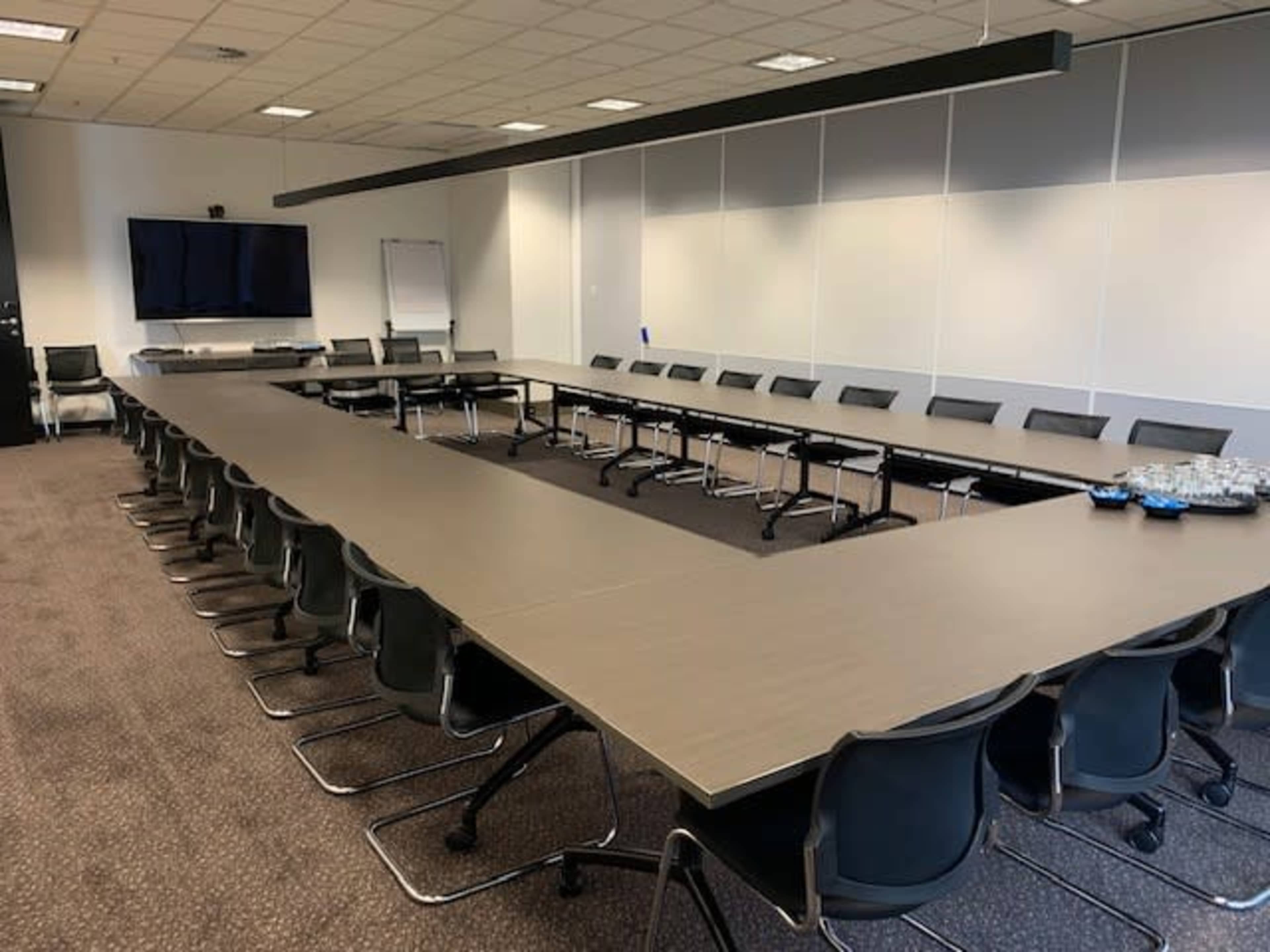The image shows a large conference room with a rectangular table surrounded by black chairs and a wall-mounted television.