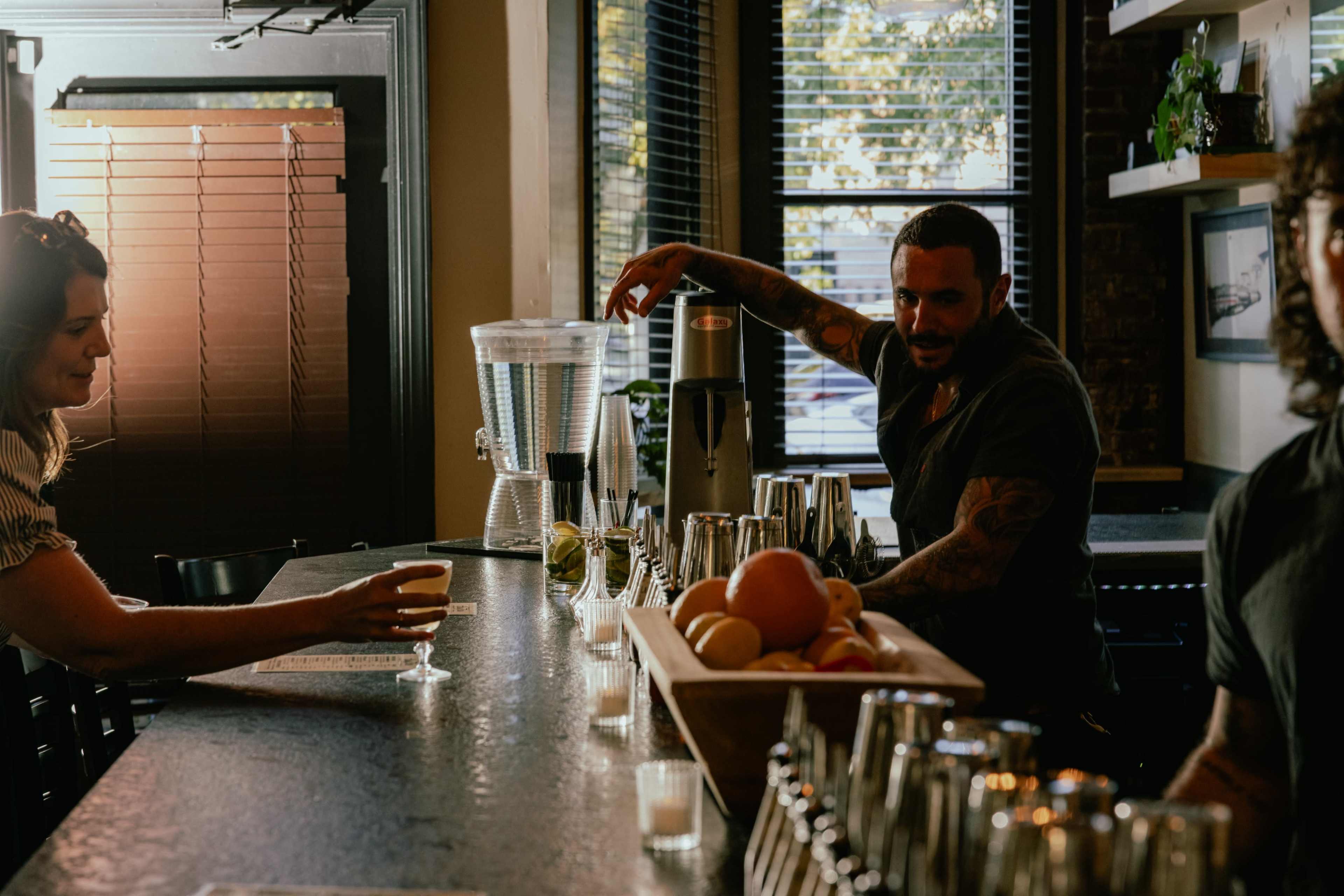 A bartender prepares a drink while a customer observes at a bar with various bottles and fruit on the counter.