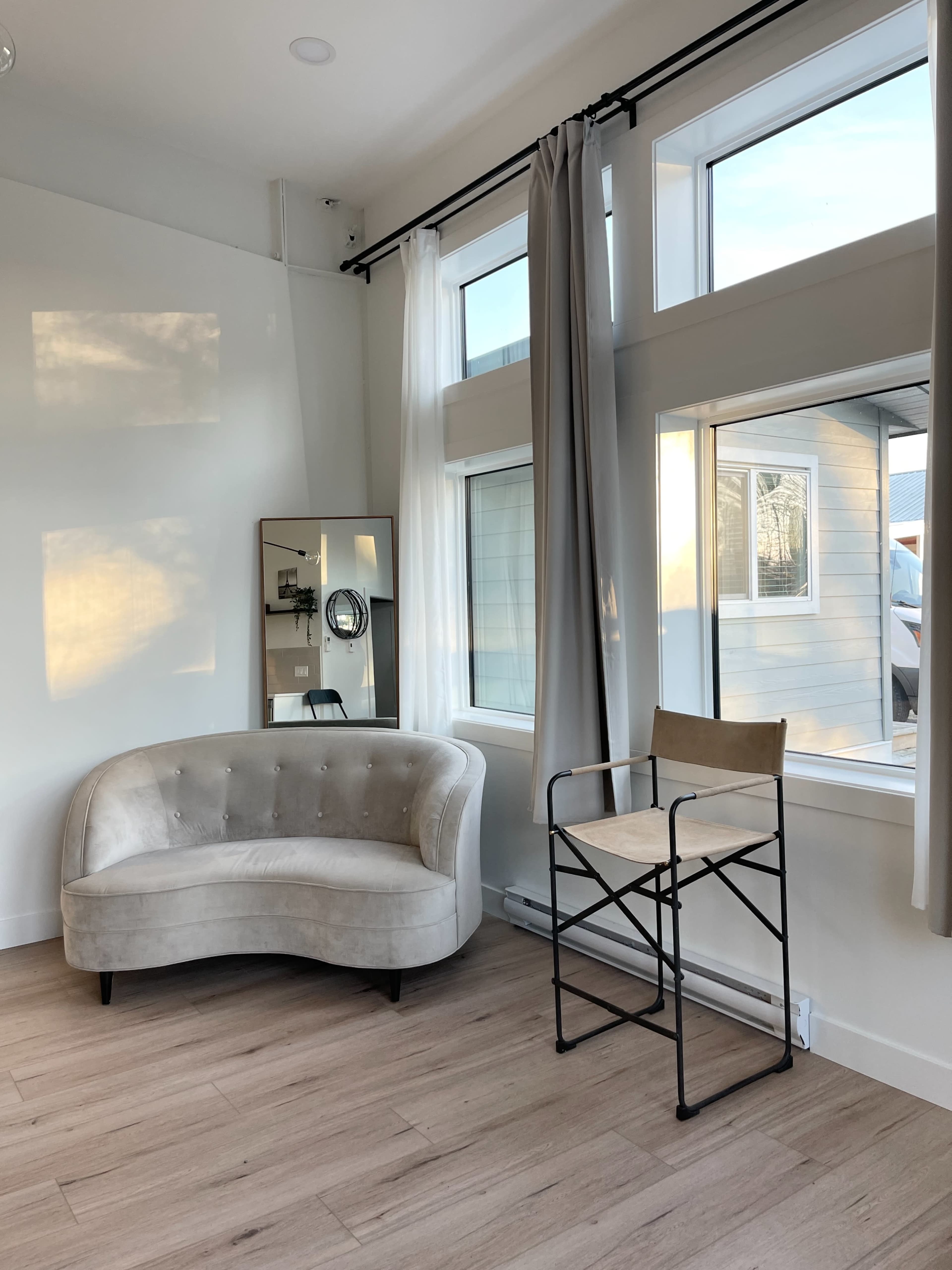 A light-filled room with a curved sofa, a tall mirror, and a folding chair beside large windows with sheer curtains.