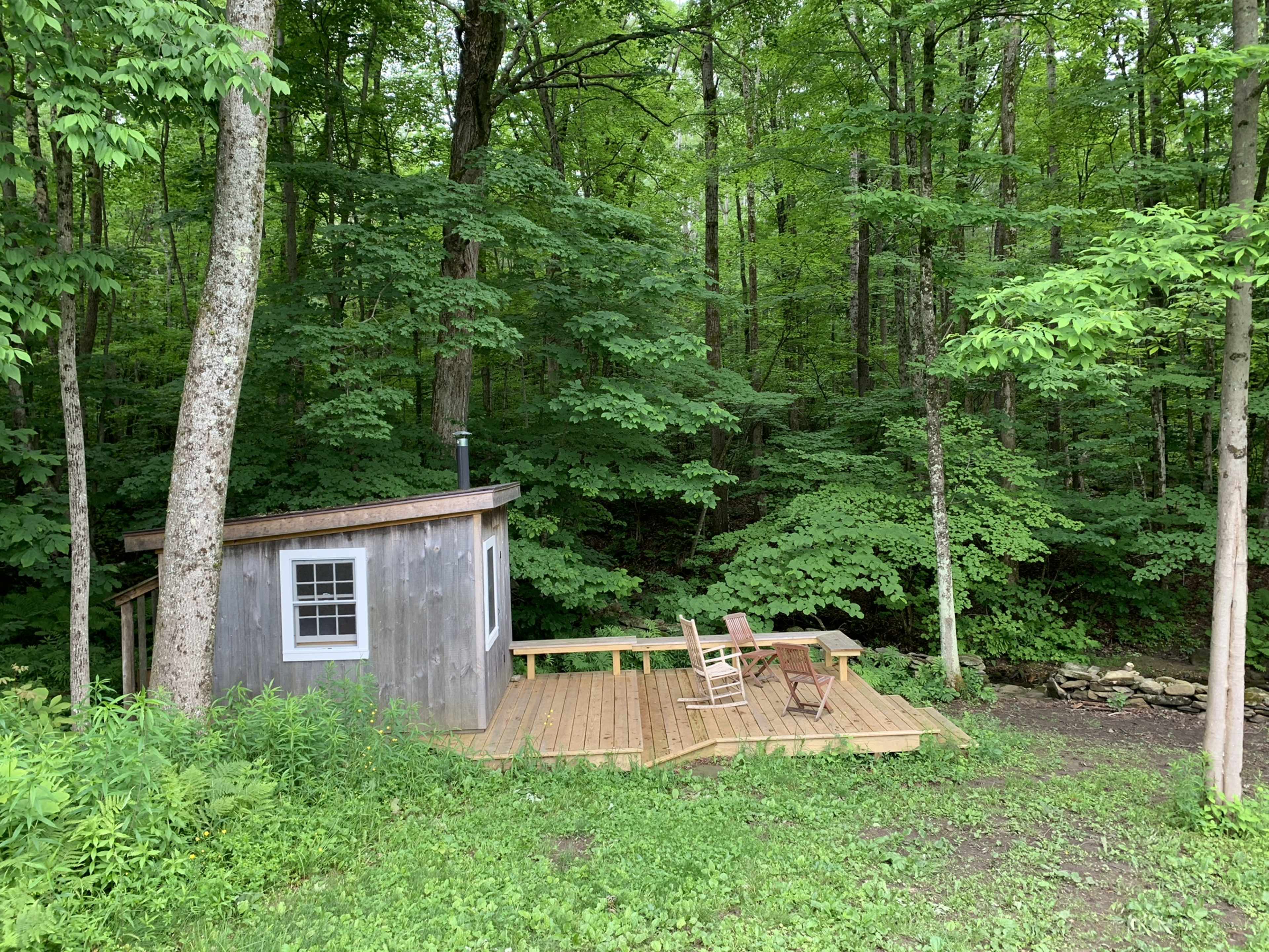 A small wooden cabin with a deck is situated among dense greenery in a wooded area.