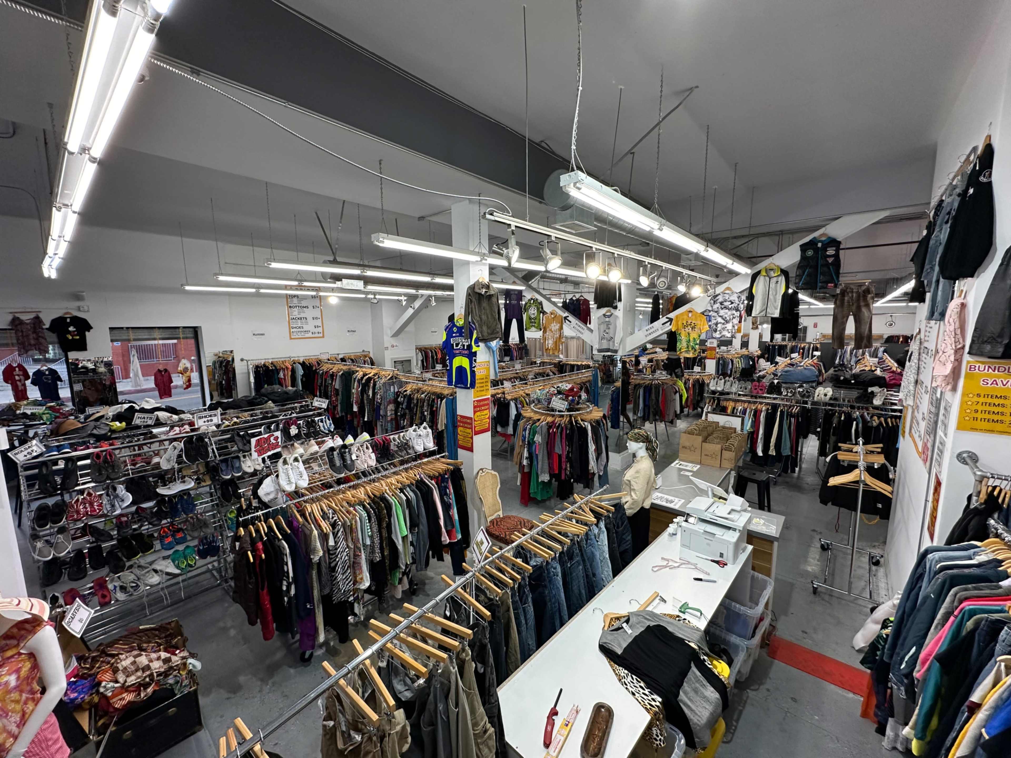 The image shows an indoor thrift store or clothing shop filled with clothing racks, organized with various garments, and multiple fluorescent lights overhead.