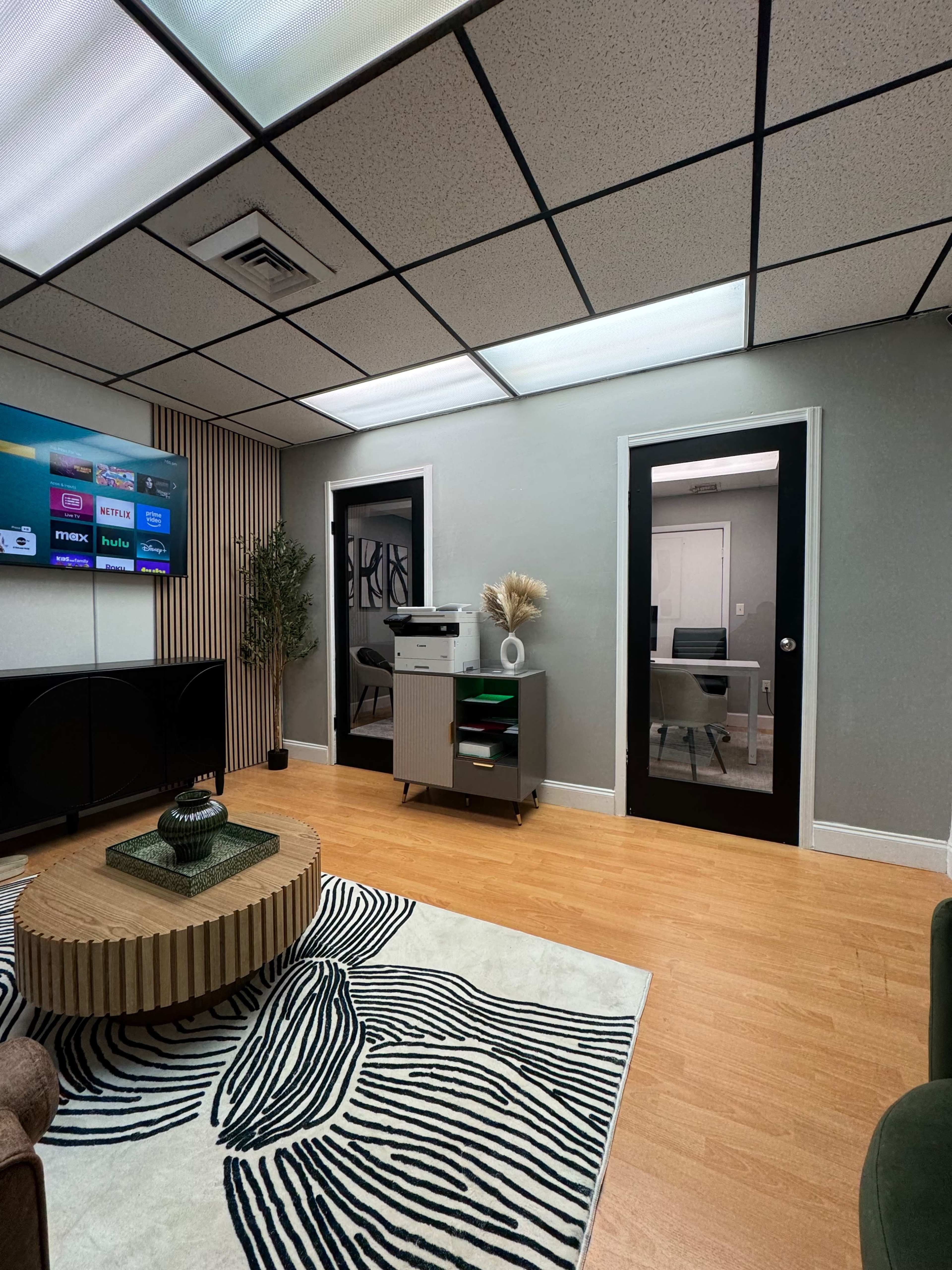 The image shows a modern office space featuring a large television on the wall, a stylish round coffee table, and two black doors leading to separate rooms.