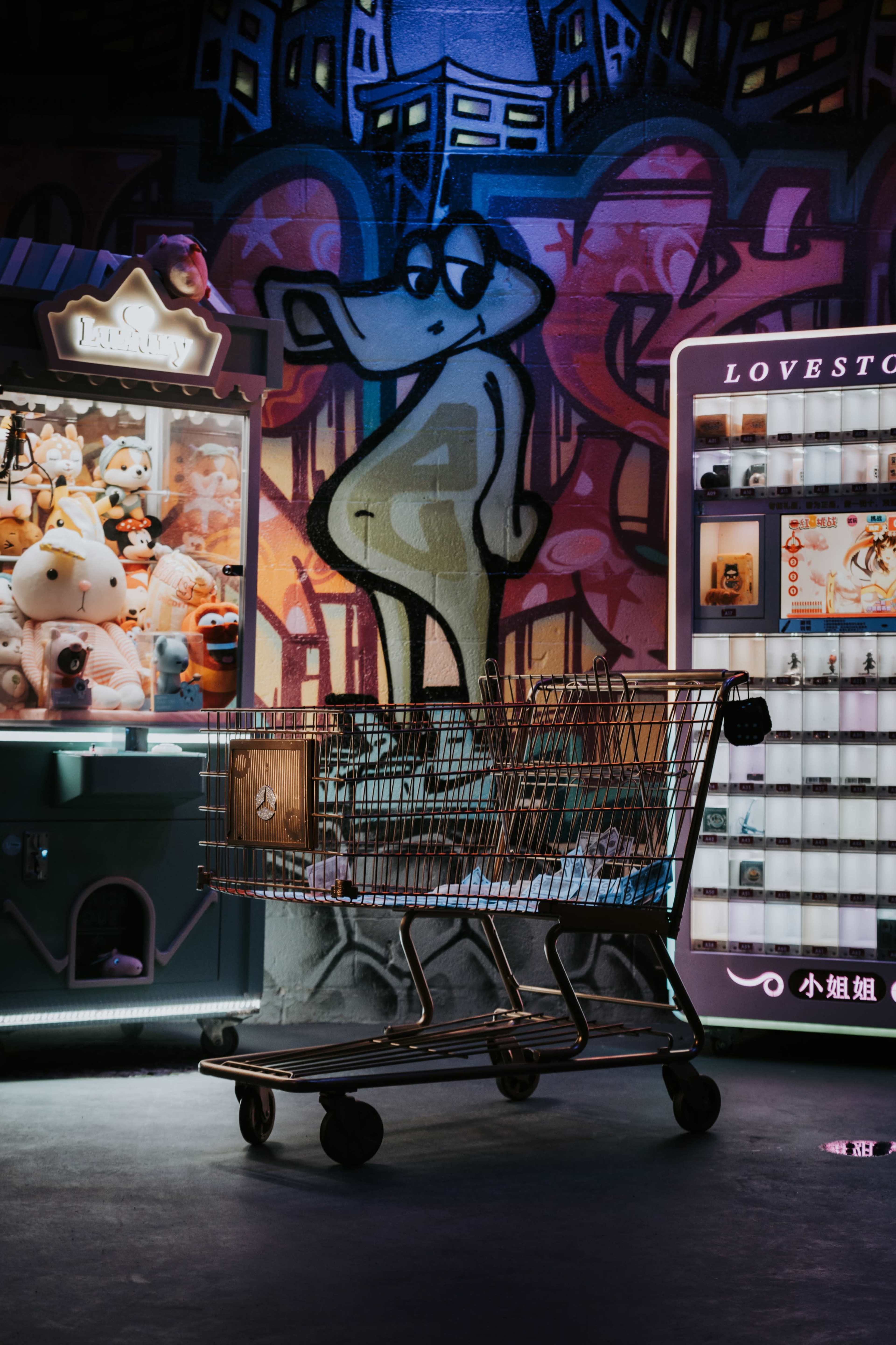 A metal shopping cart is centered in front of a colorful graffiti wall and two vending machines filled with toys and trinkets.
