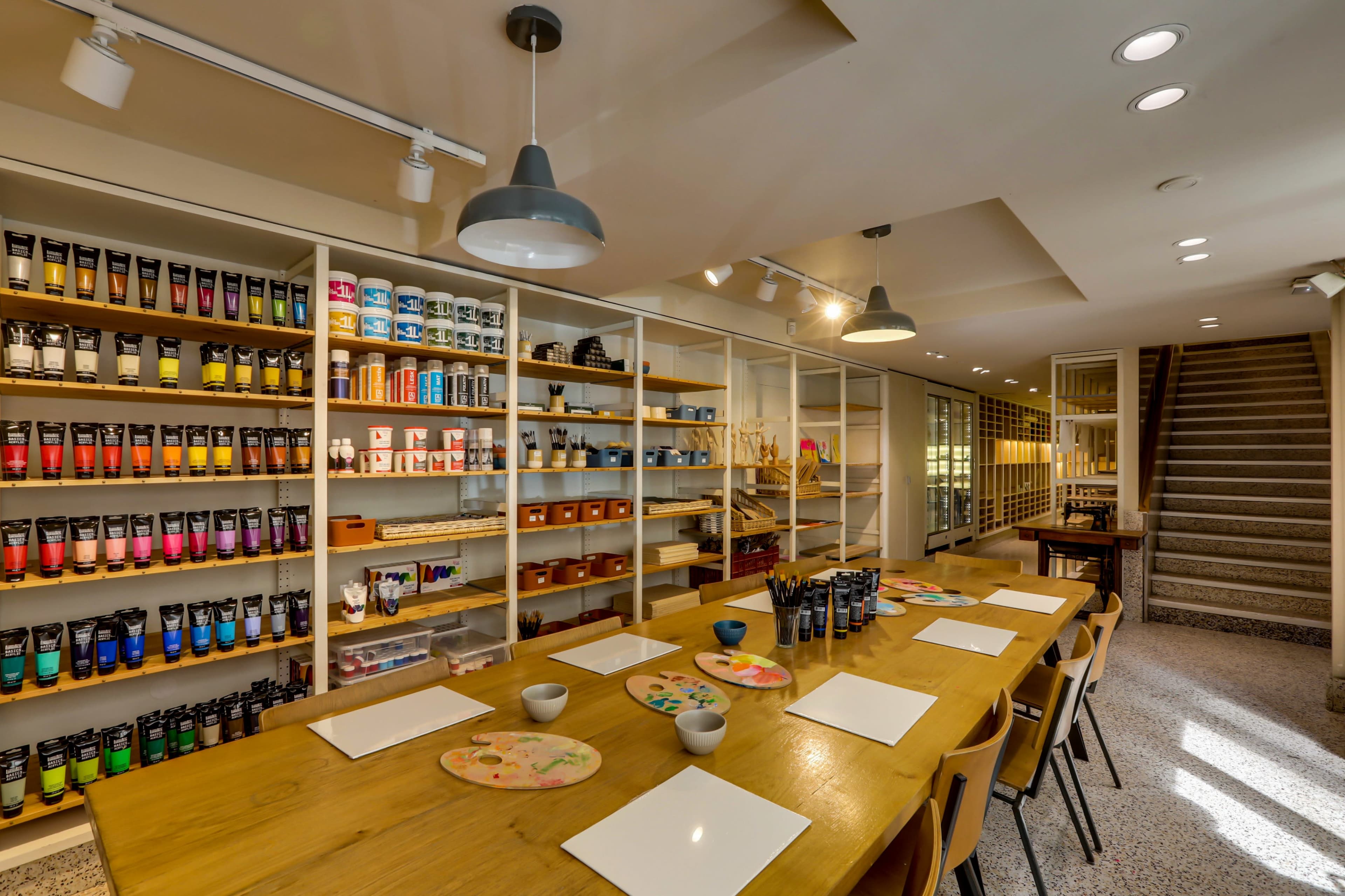 The image shows a well-lit art studio with shelves of paint and supplies arranged neatly alongside a large table set with blank canvases and art materials.