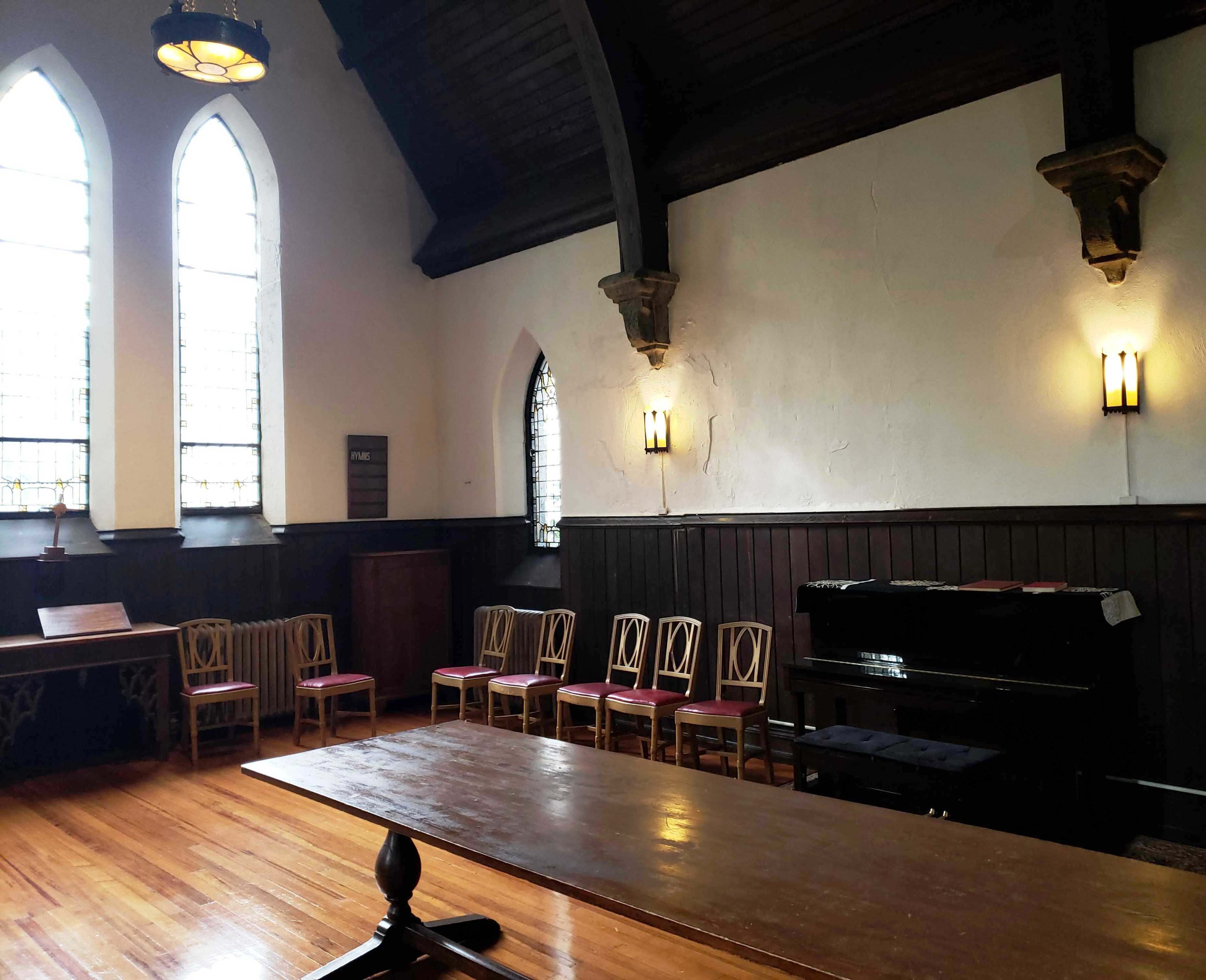 The interior of a church features a wooden table in the foreground, seven chairs arranged along one wall, and a black piano against another wall, illuminated by light from stained glass windows.